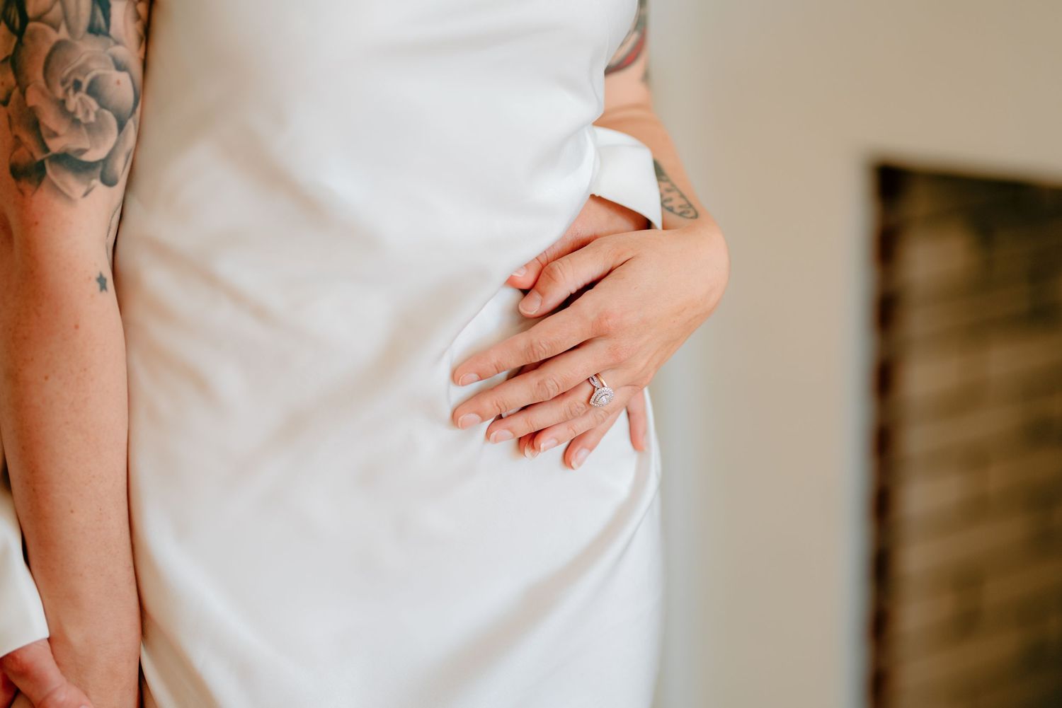 Bride in white wedding gown displaying diamond engagement ring, with visible tattoos on arm.