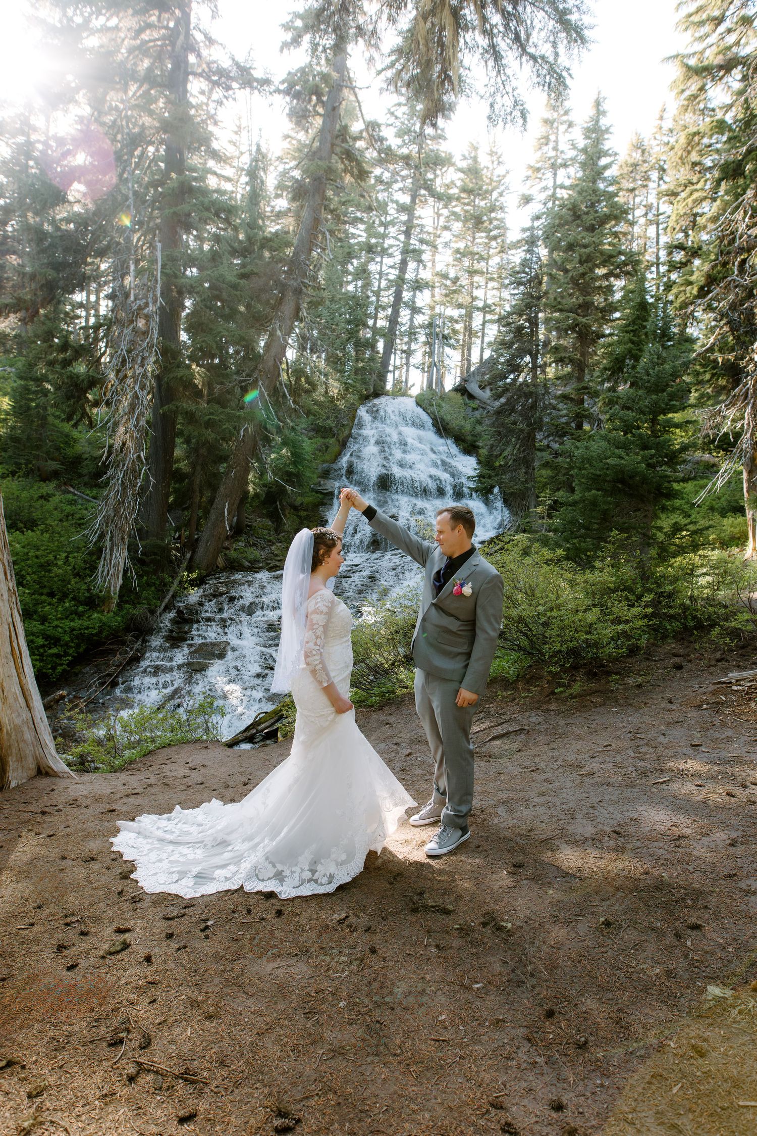 Bride and groom dance near a forest waterfall, bride in lace gown with veil, groom in gray suit, surrounded by tall pines.