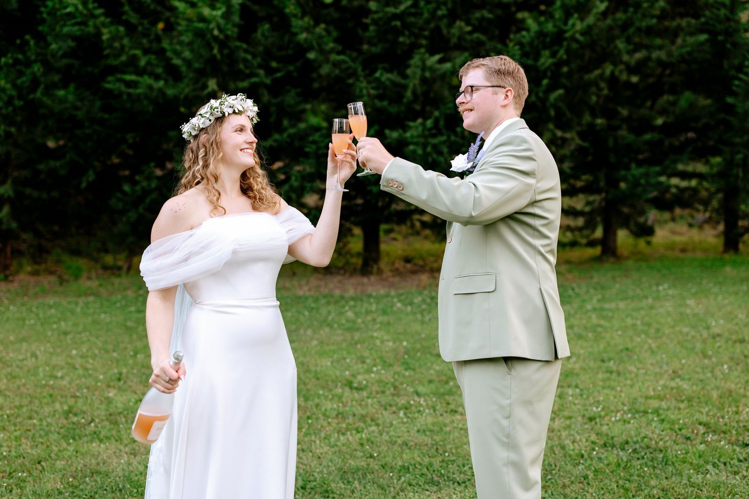 Bride in white gown with floral crown and groom in sage suit toasting champagne outdoors.