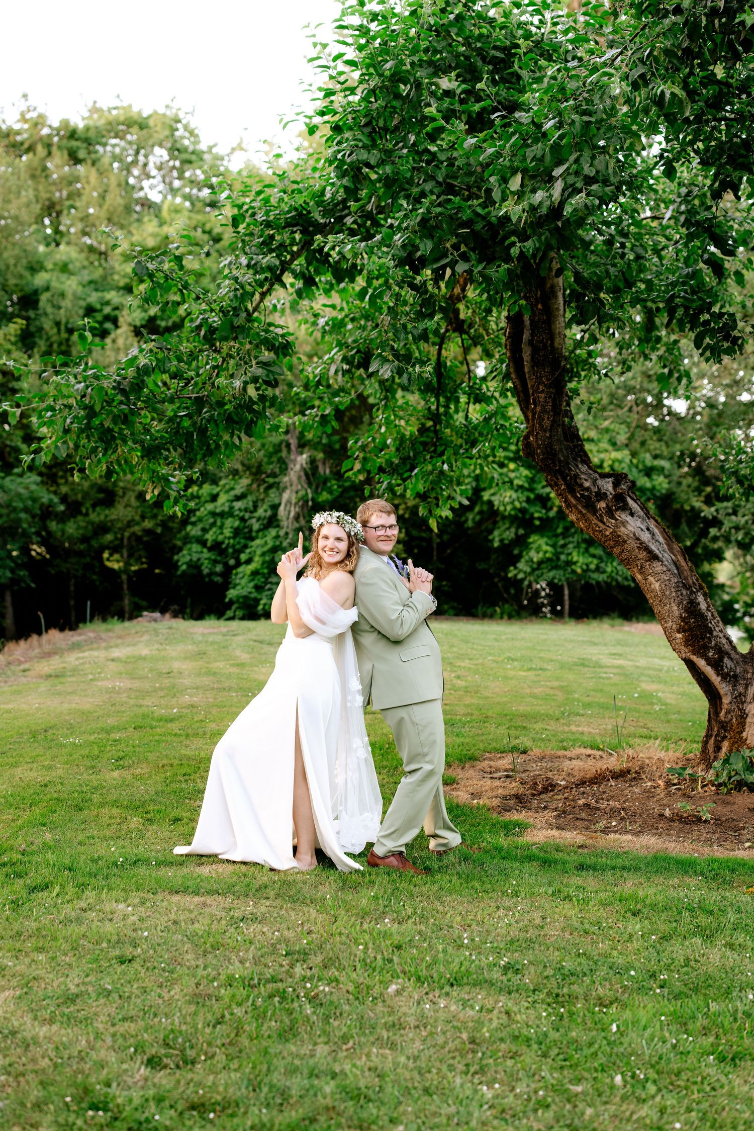 Bride in floral crown and groom in sage suit embrace while posing beside a lush green tree.