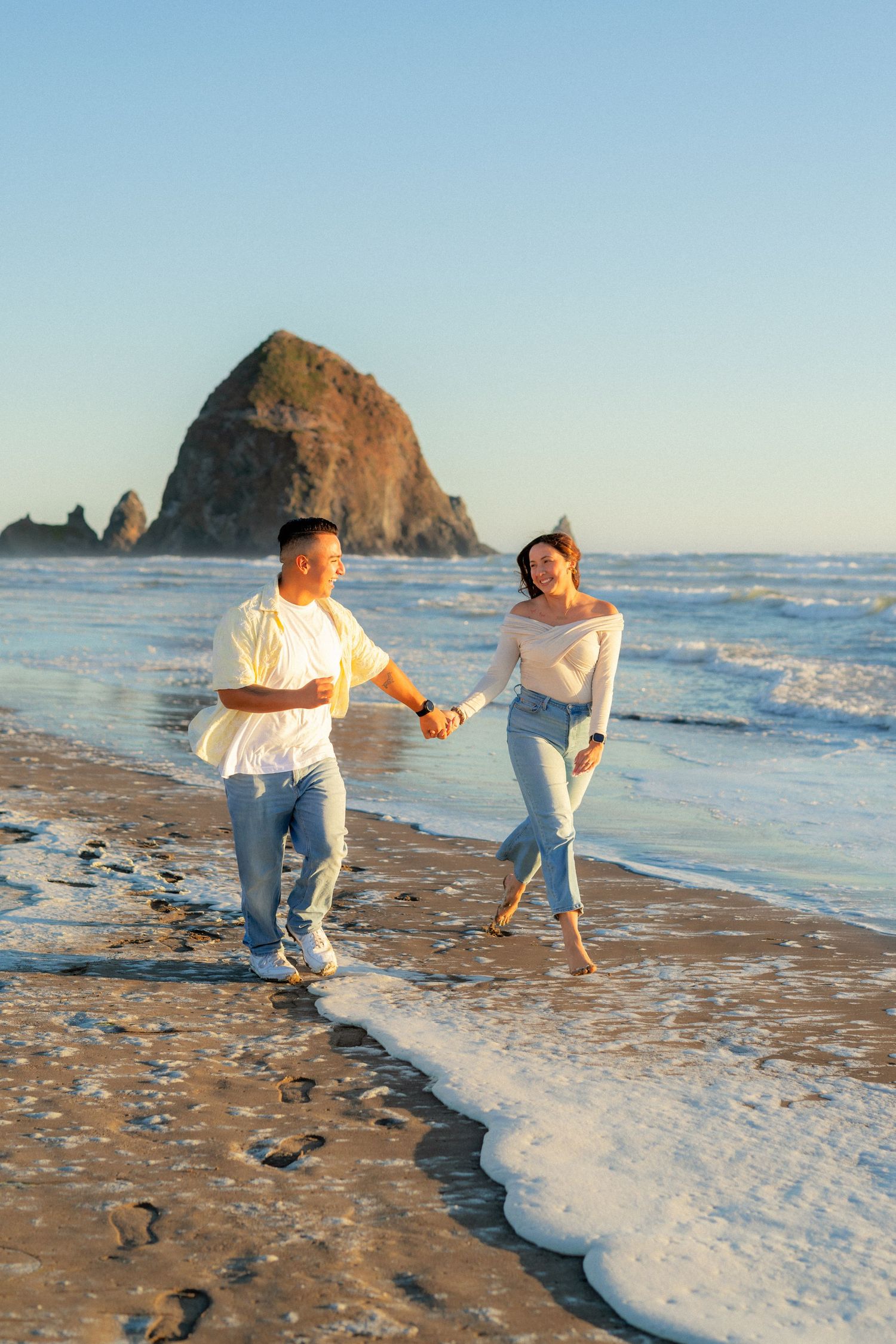 Happy couple holding hands running along Cannon Beach shoreline with Haystack Rock behind them.