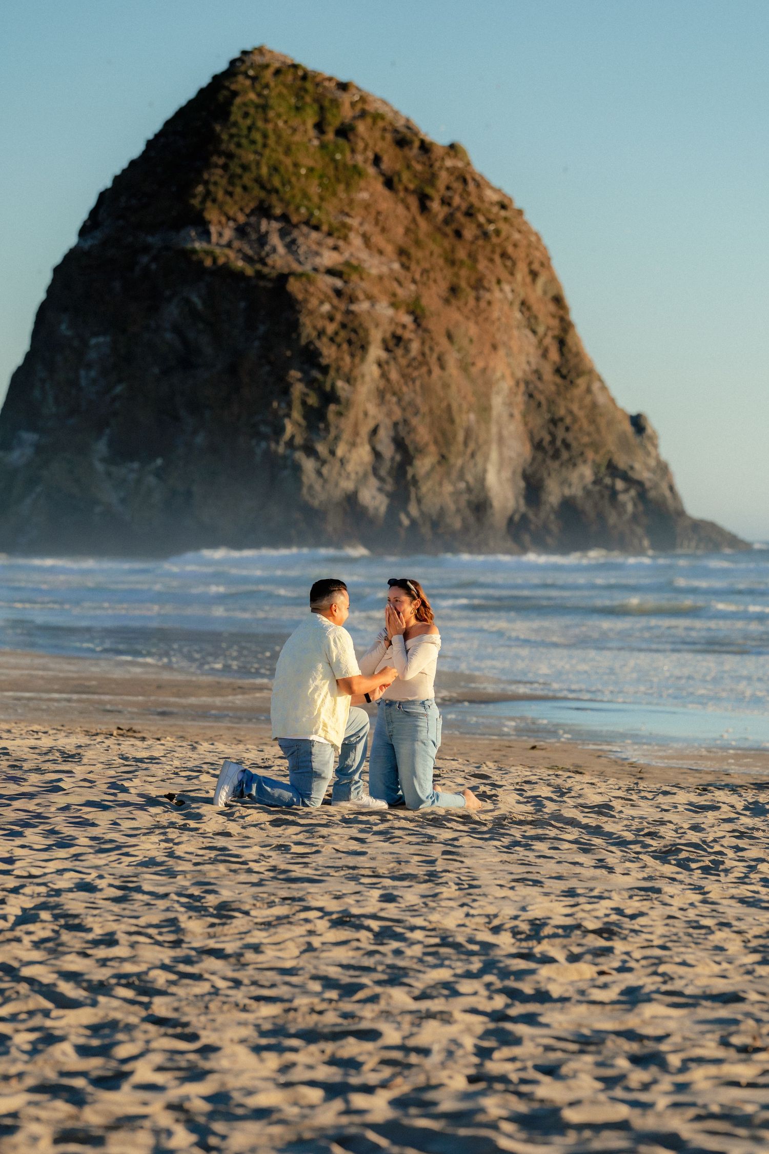 Couple sharing a romantic beach proposal moment on sandy shore with iconic Haystack Rock at Cannon Beach, Oregon backdrop.