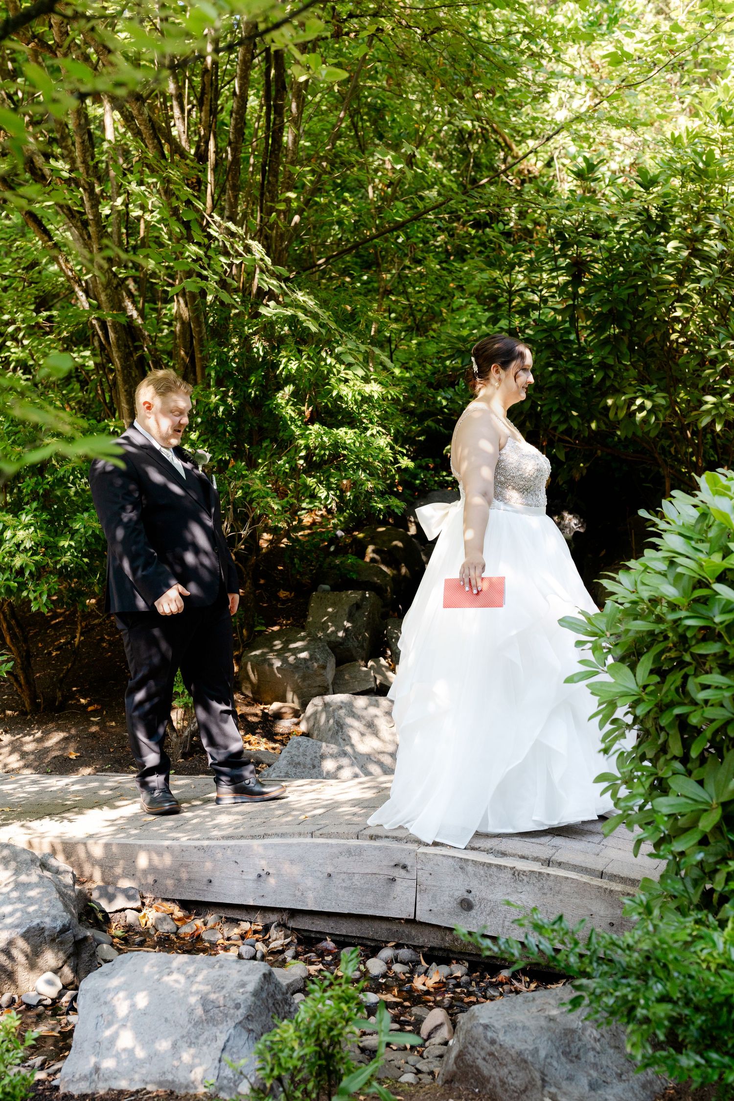 Bride in white gown and groom in black suit walk along stone path surrounded by lush green trees.