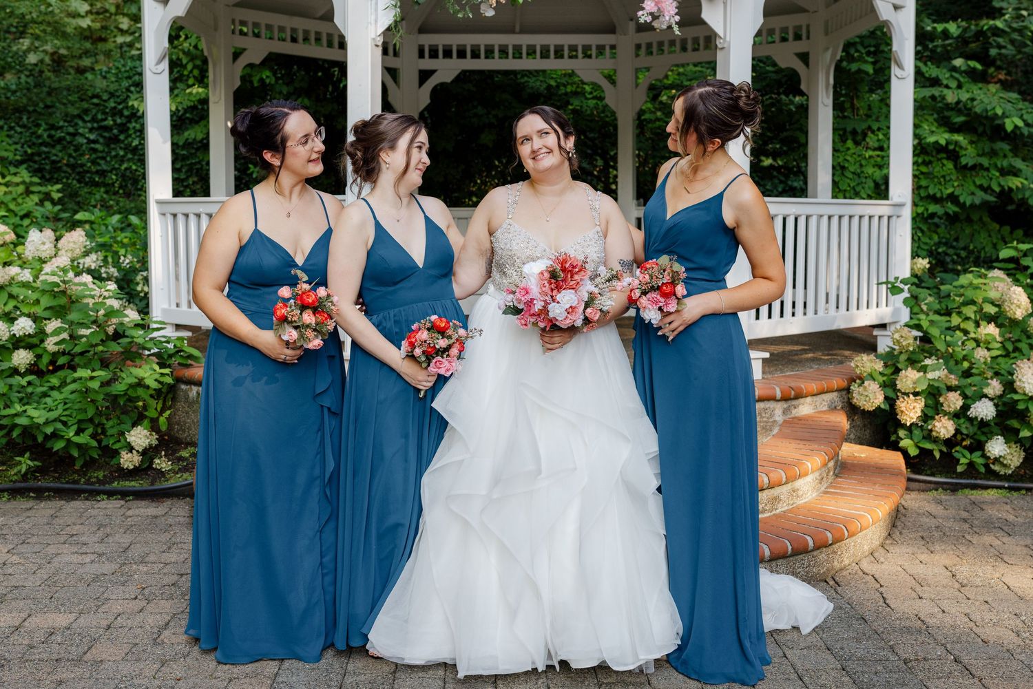 Bride poses with three bridesmaids in blue dresses holding colorful bouquets near white gazebo.