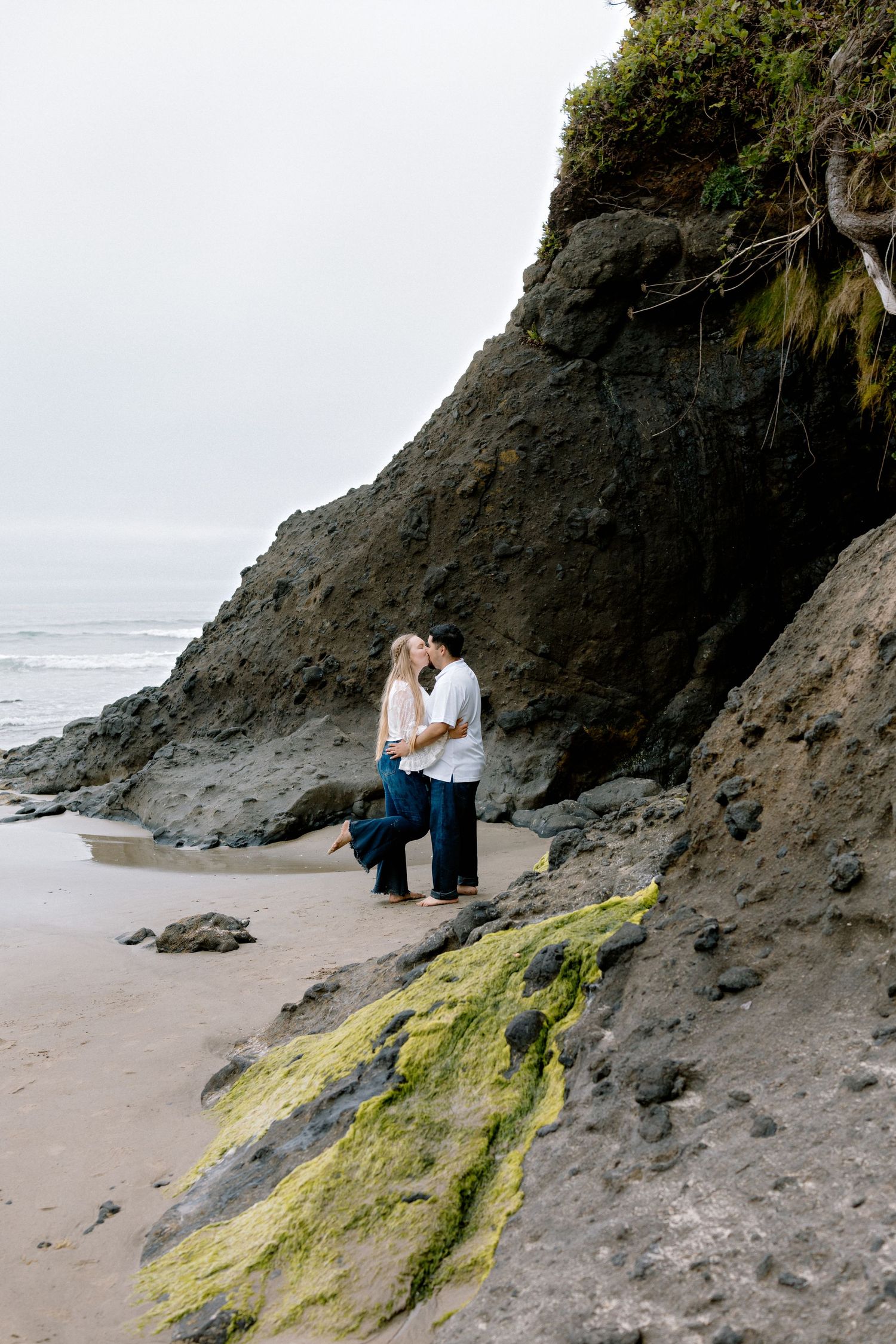 Couple sharing a romantic kiss near dramatic rocky cliffs on a misty Pacific Northwest beach.