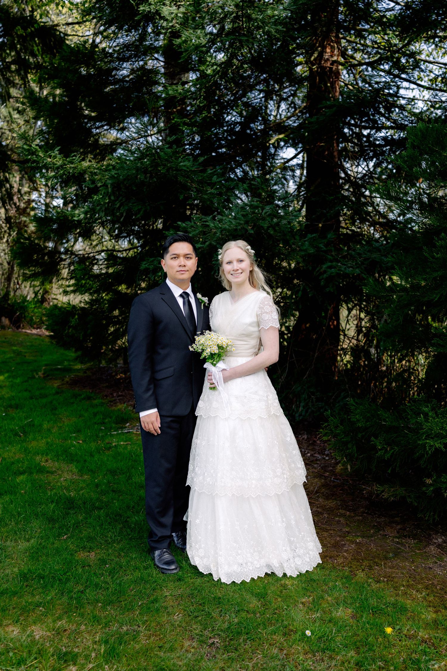 Bride and groom posing together in formal wedding attire in front of evergreen trees.