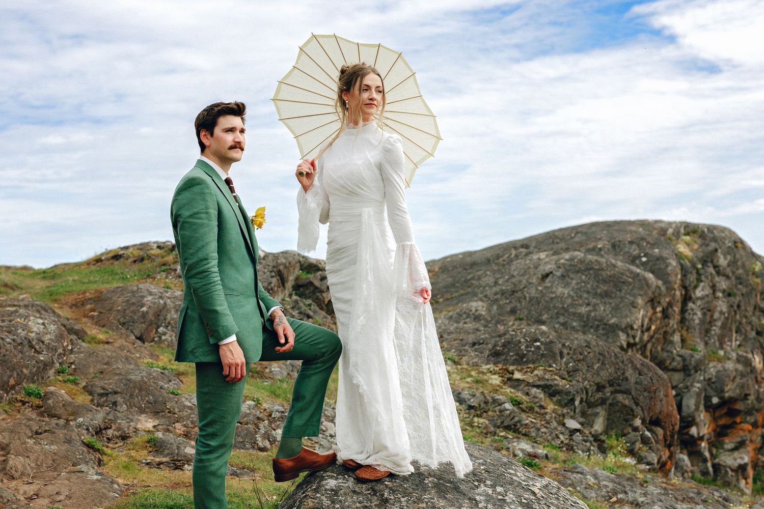 Stylish wedding couple on rocky terrain, groom in green suit with yellow flower, bride in white lace dress holding parasol.