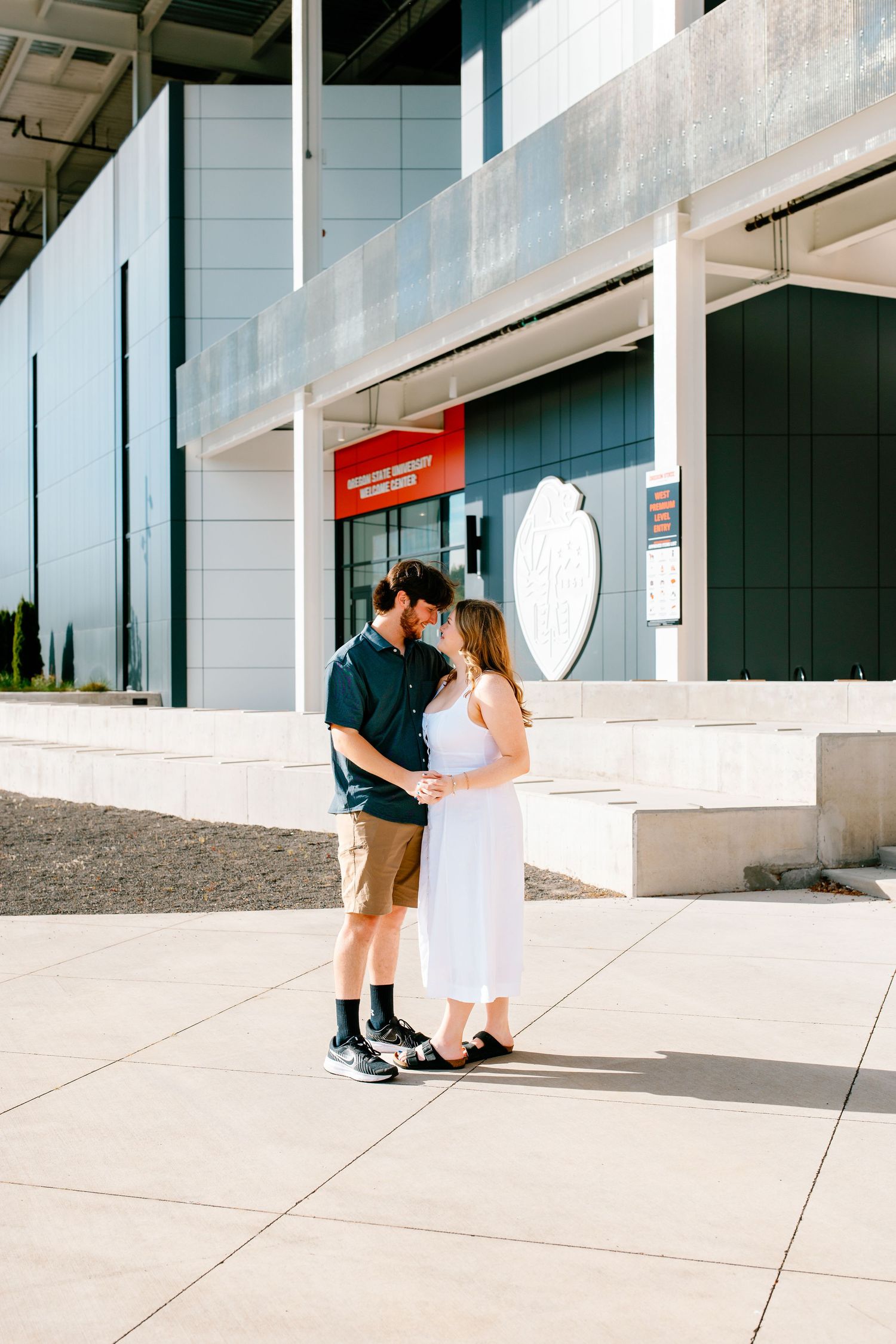 A couple shares a romantic moment outside a modern building, with the woman in a white dress and man in casual attire.