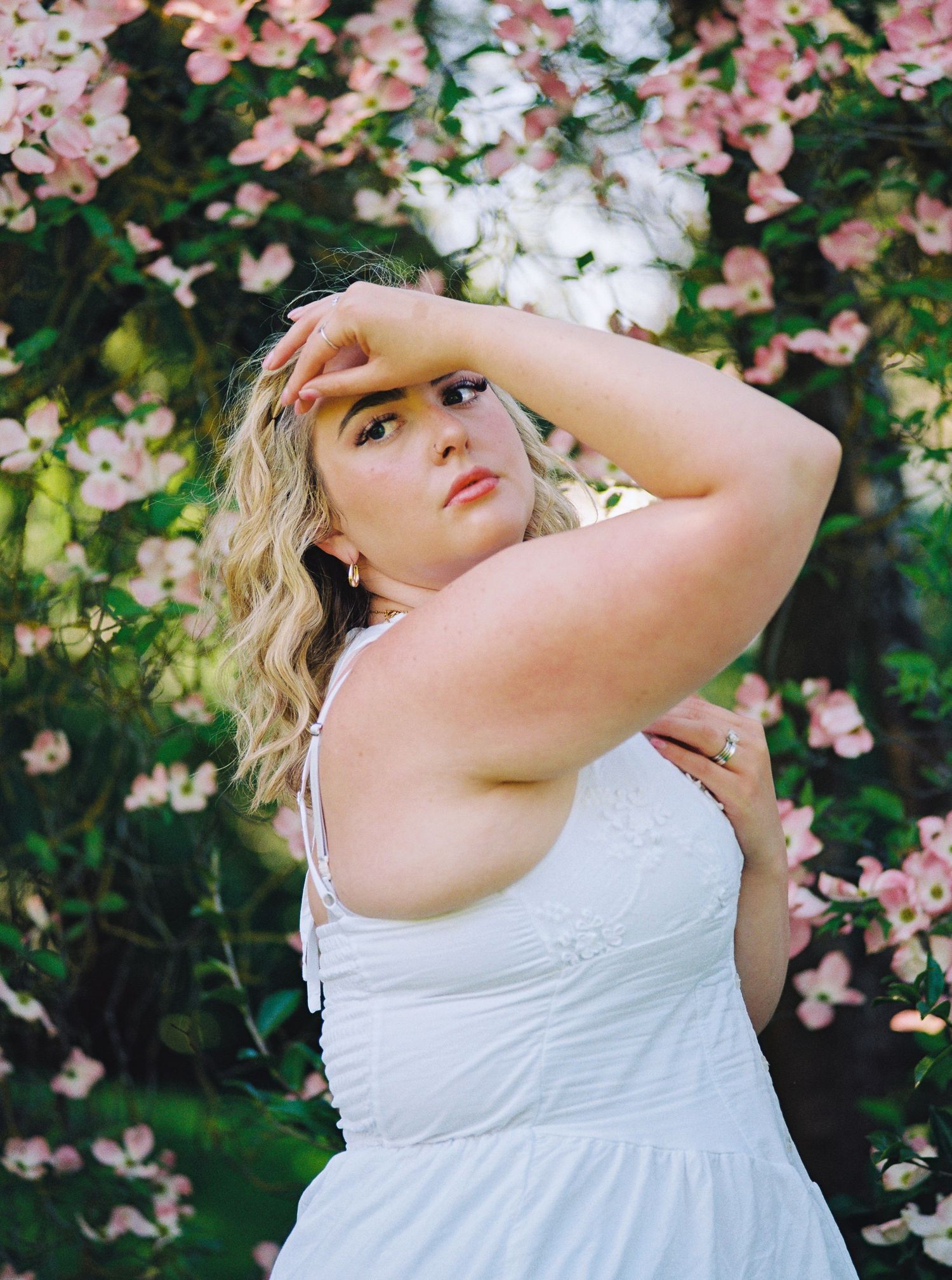 Blonde woman in white sundress posing among pink flowering dogwood trees in garden setting.