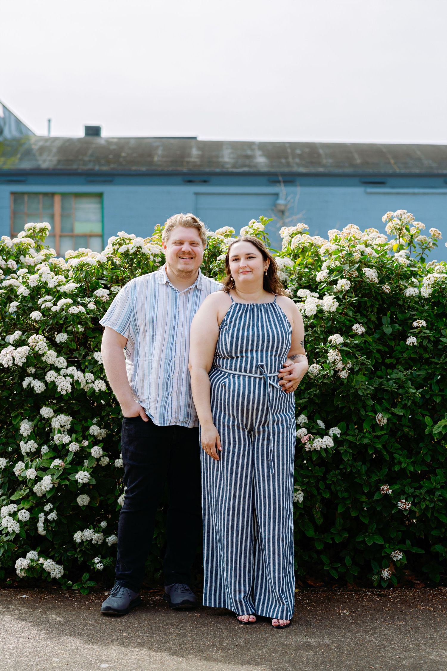 Two people standing together in front of a hedge with white blooming flowers on a sunny day.