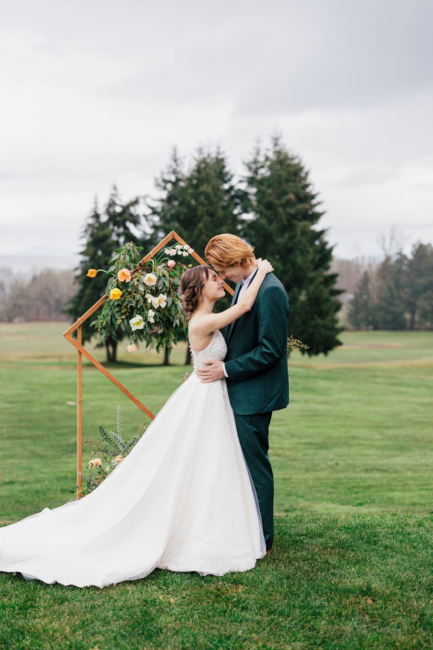 A couple shares a romantic moment under a floral arch on a misty green lawn with evergreen trees in the background.