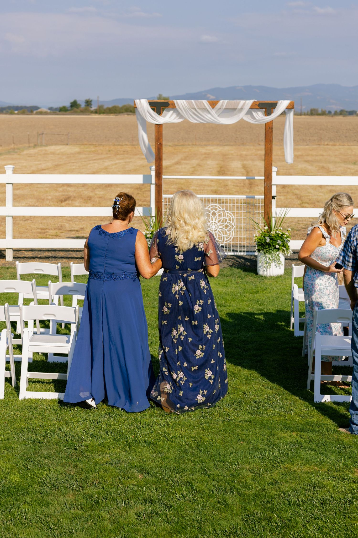 Outdoor wedding ceremony setup with white chairs and floral arrangements on a grassy lawn with mountains in the background.