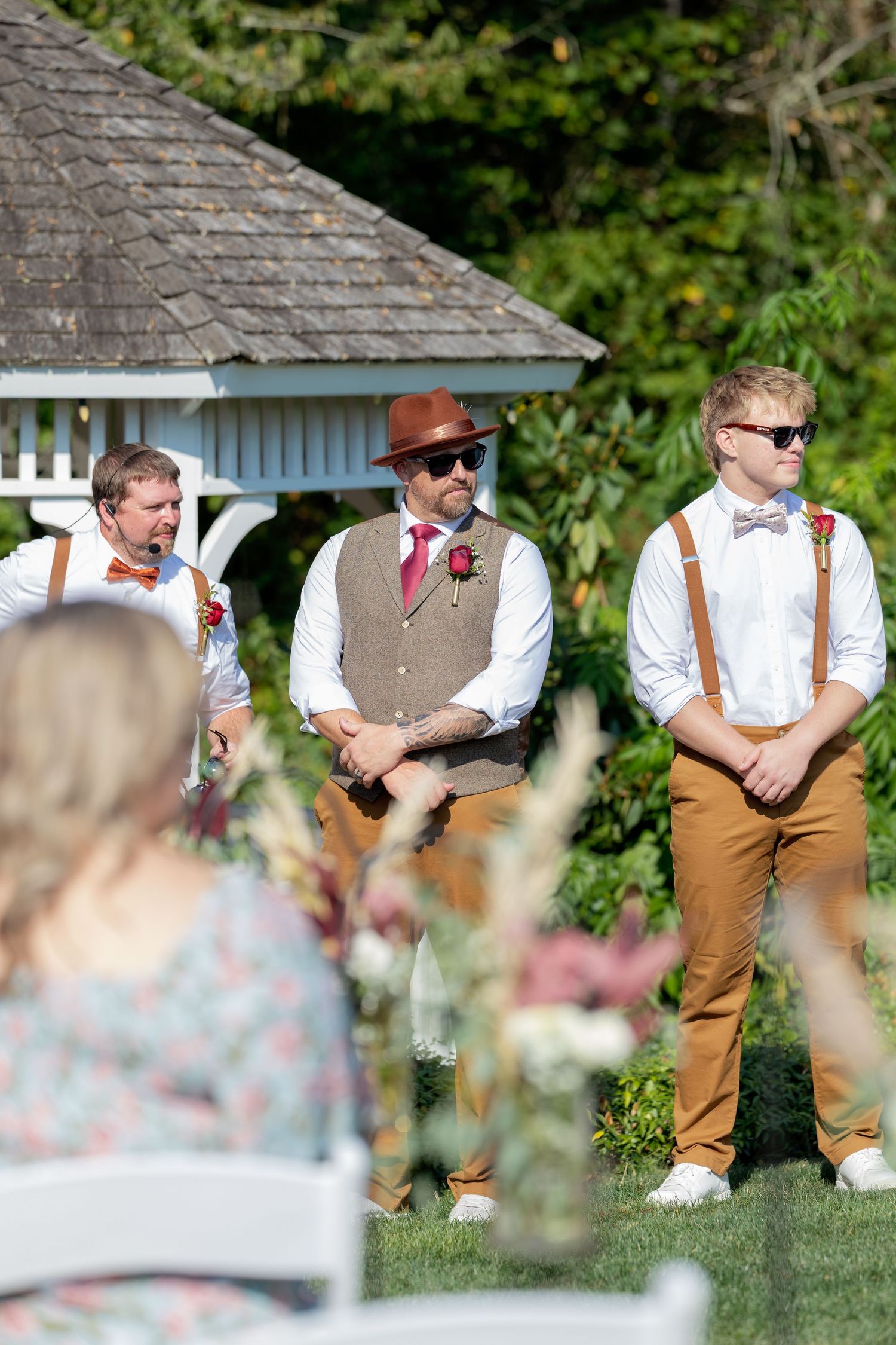 Wedding party members in brown vests and suspenders sitting casually on a stone wall outdoors.