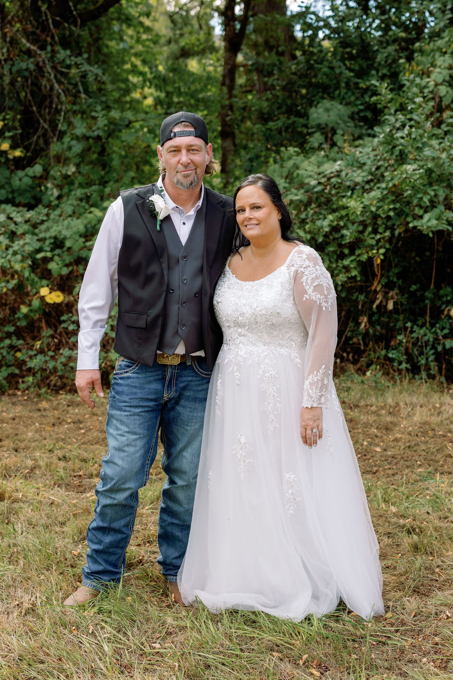 A wedding couple poses outdoors in casual country attire with the bride in a lace gown and groom in jeans and vest.