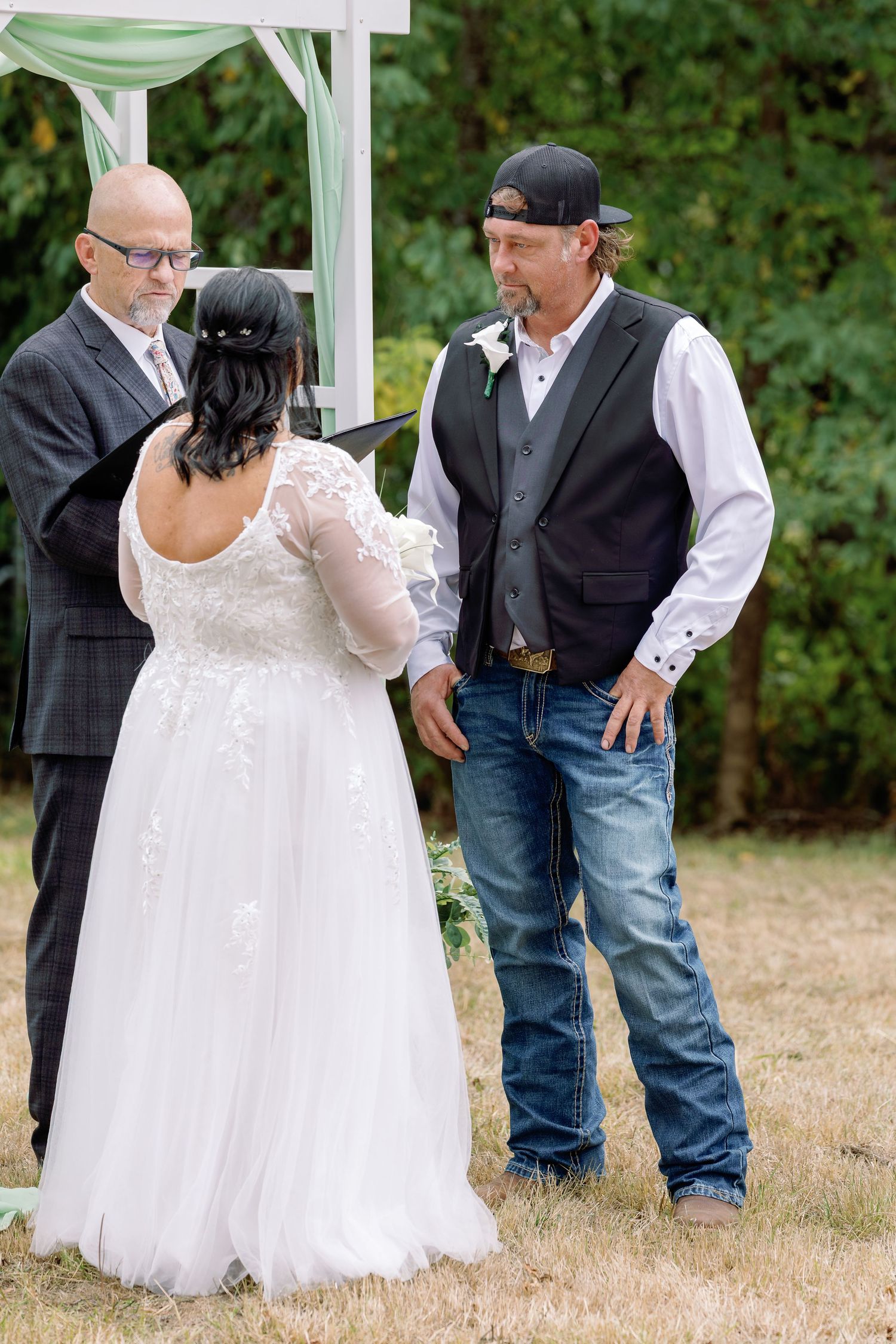 A wedding ceremony takes place outdoors under a white canopy with the officiant and couple in casual formal attire.