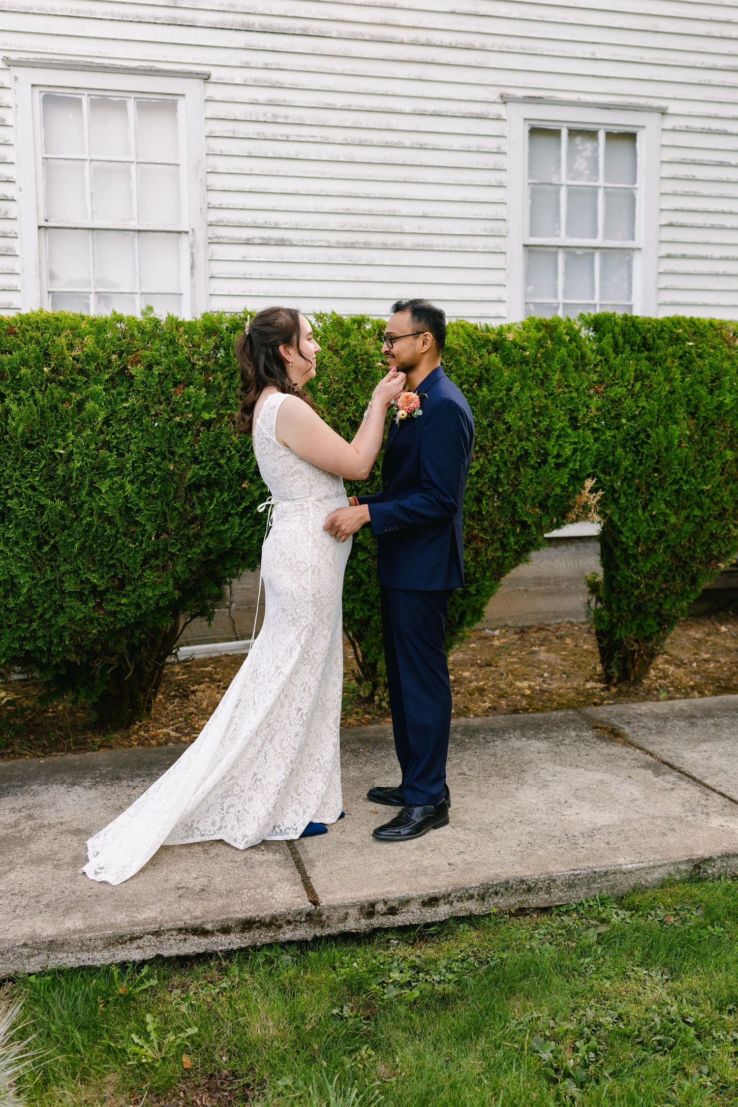 A couple in wedding attire dances playfully in front of a white house with green hedges.