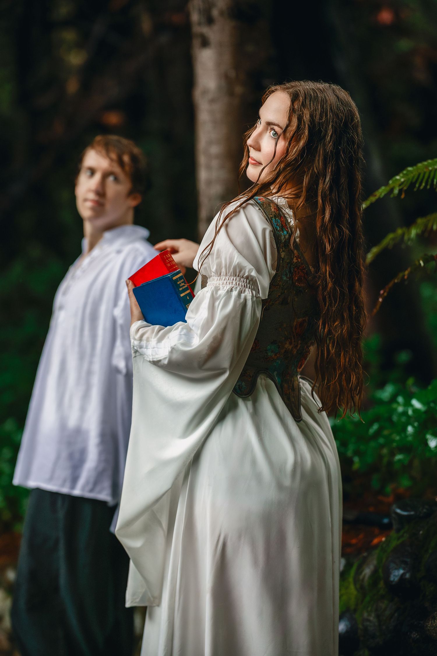 A couple in white period clothing poses in a dreamy forest setting while holding a book.