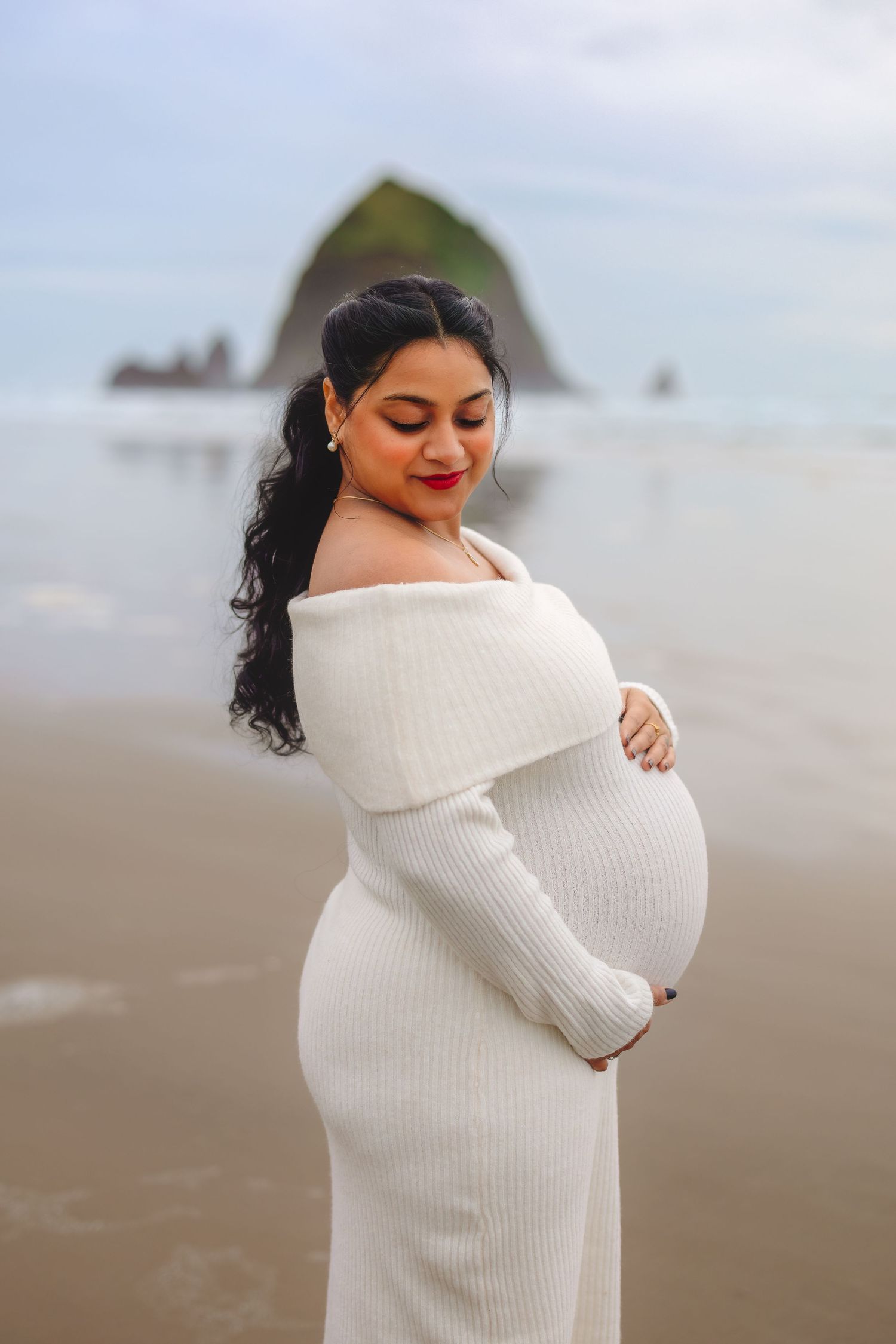 Expectant mother in white sweater dress poses on beach with dramatic coastal landscape behind her.