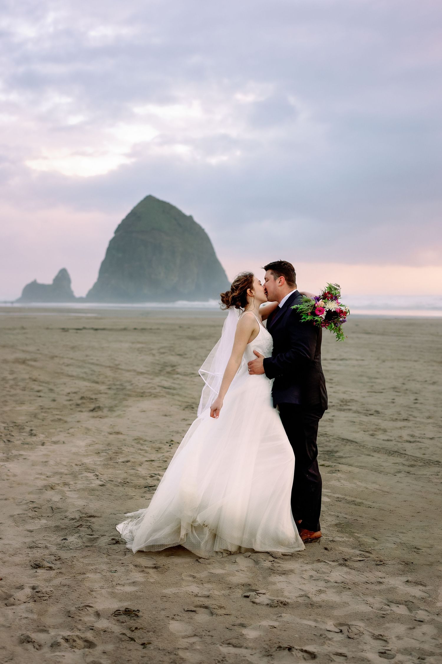 A romantic wedding couple embraces on a misty beach with dramatic rock formations in the background at sunset.