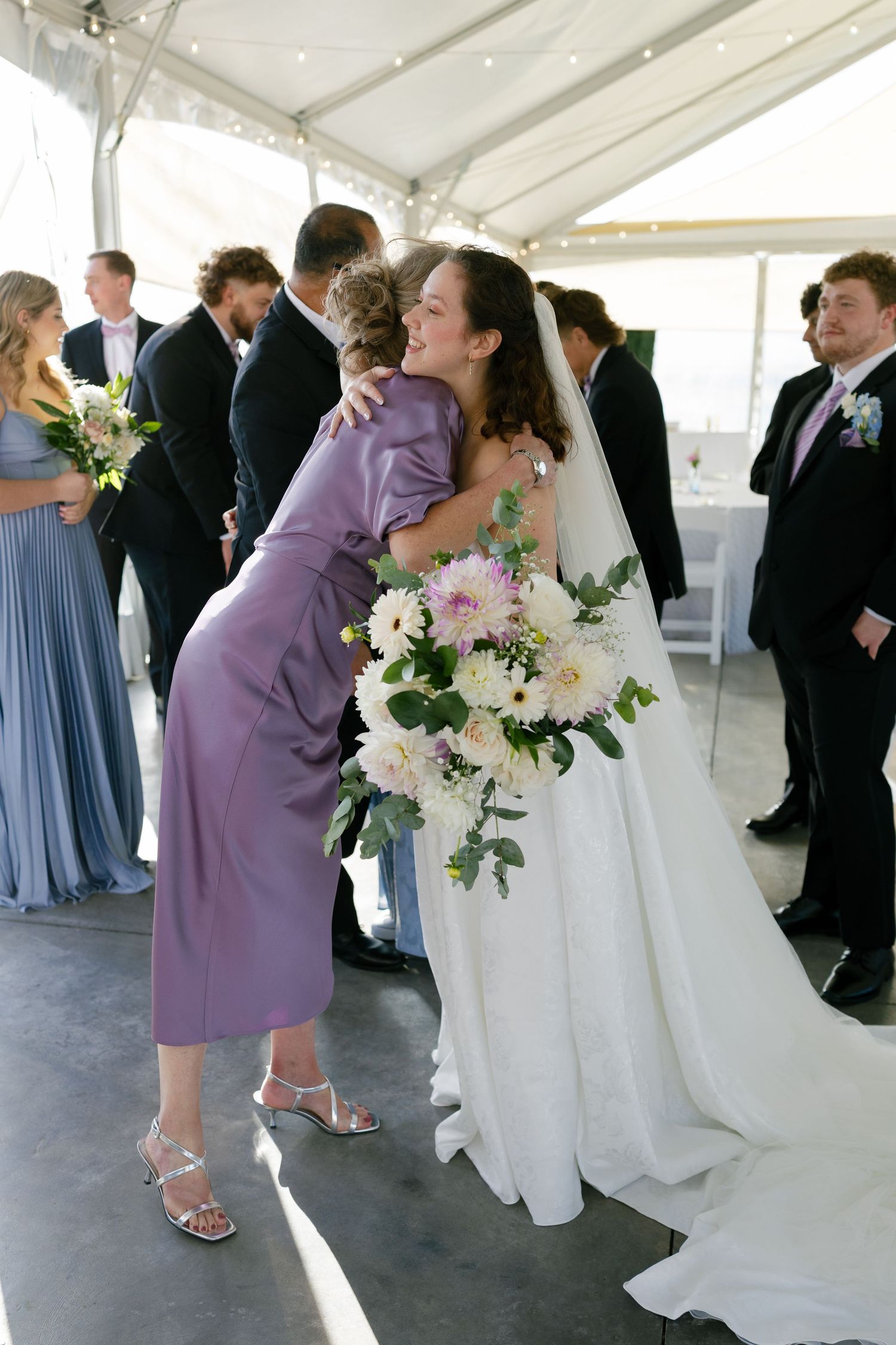 Wedding party members in formal attire with bridesmaids in light blue dresses and purple dresses under a white tent.