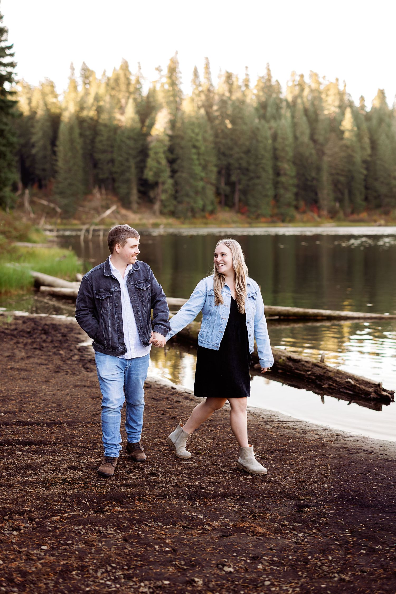 Two people strolling along a serene lakeshore with evergreen trees in the background.