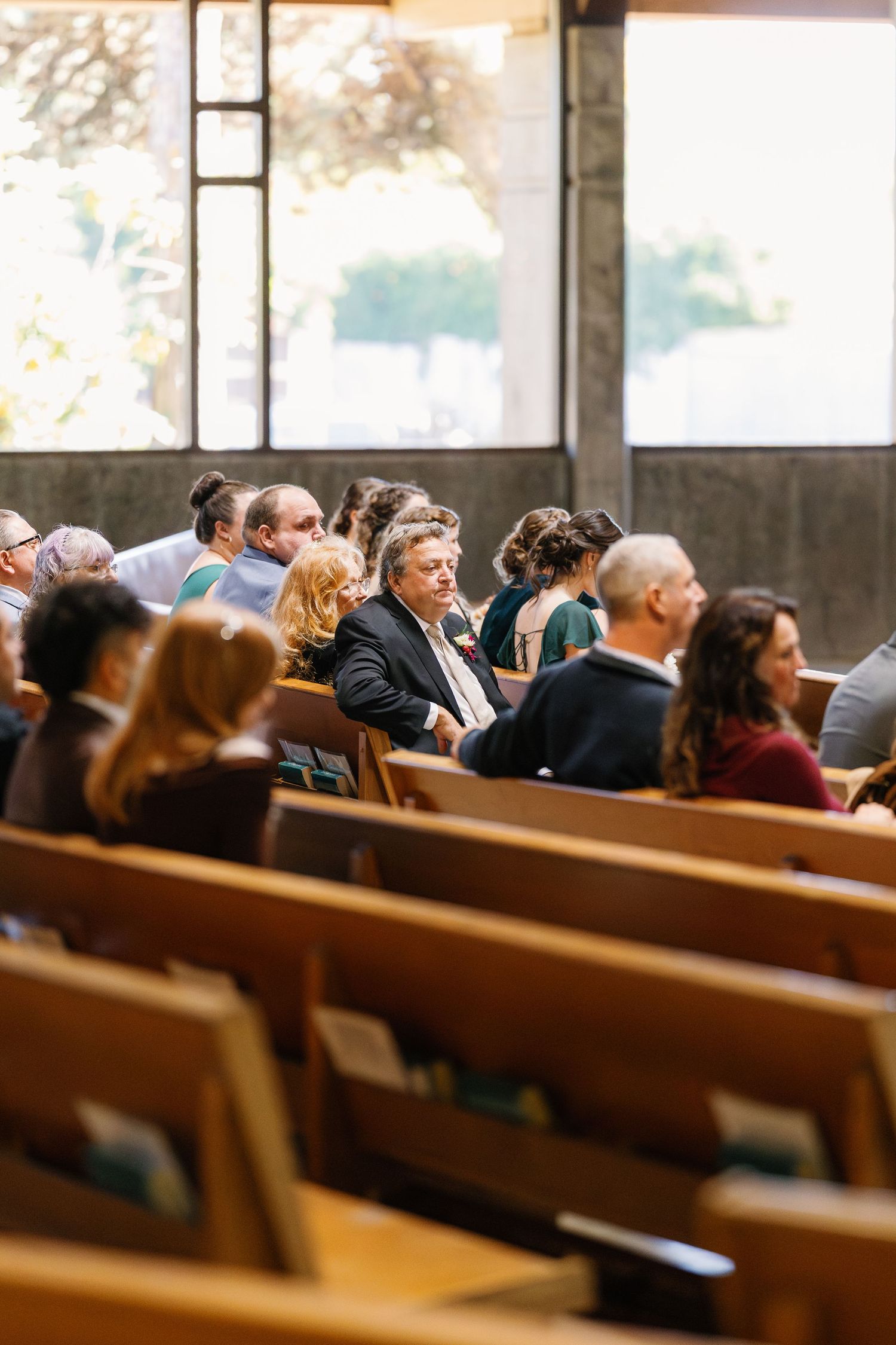Guests seated in wooden church pews during a wedding ceremony with natural light streaming through windows.