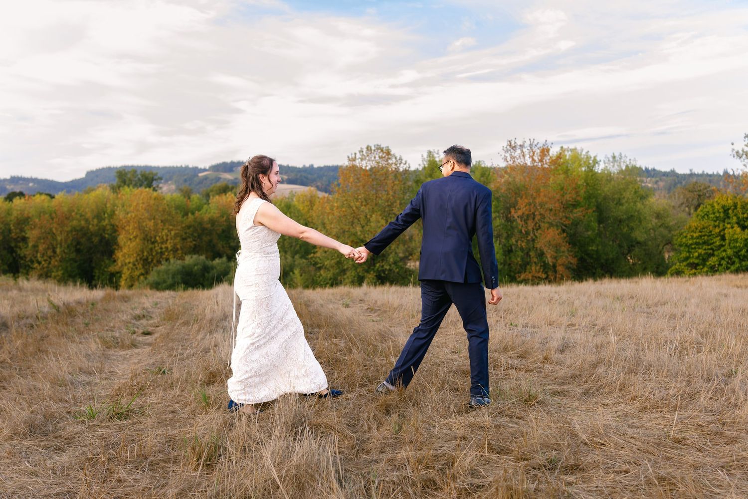 Two people in wedding attire walk together through a golden field during autumn sunset.
