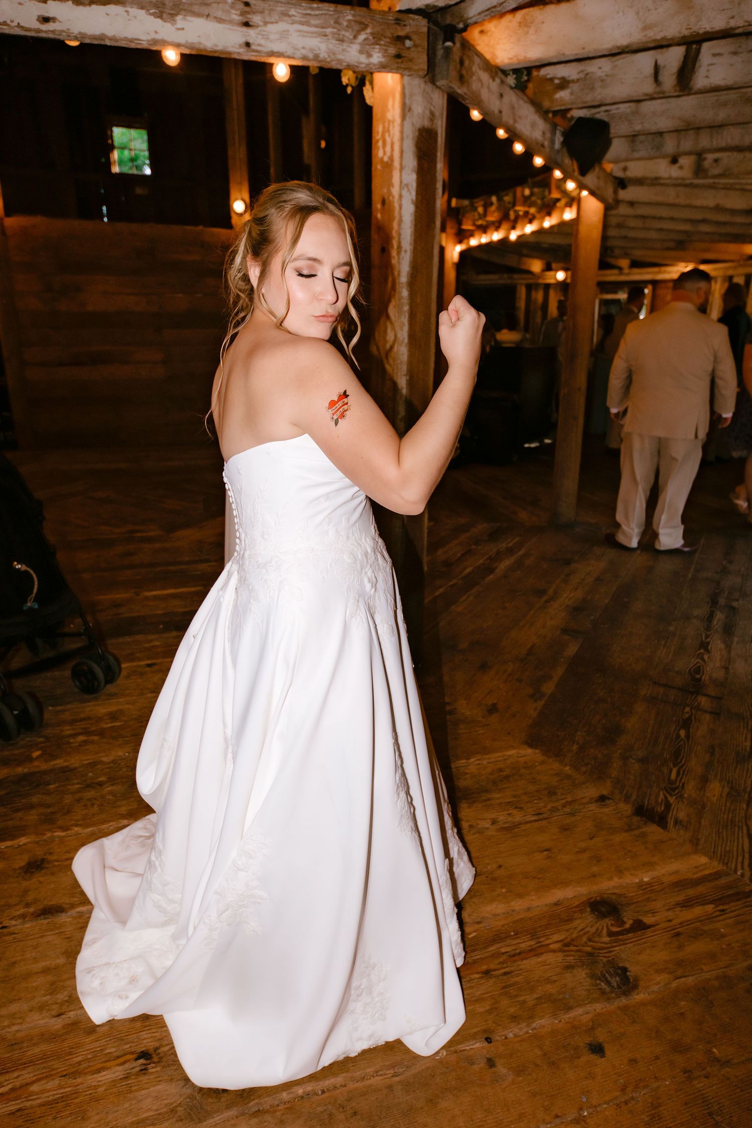 Bride in strapless white gown dancing in rustic barn venue with string lights and wooden beams.
