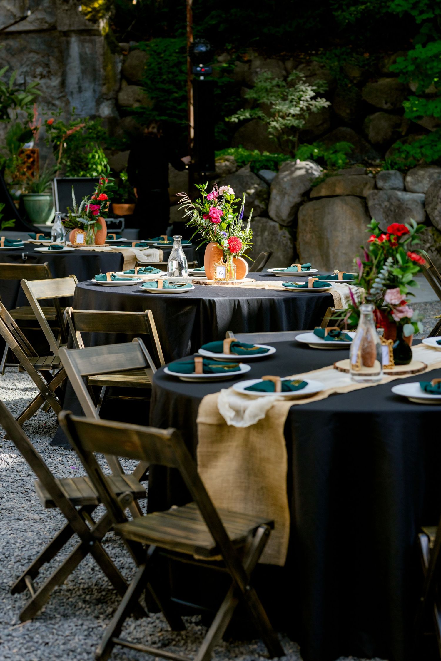Outdoor wedding reception tables with black linens, burlap runners, teal napkins, and floral centerpieces set against a stone wall backdrop.