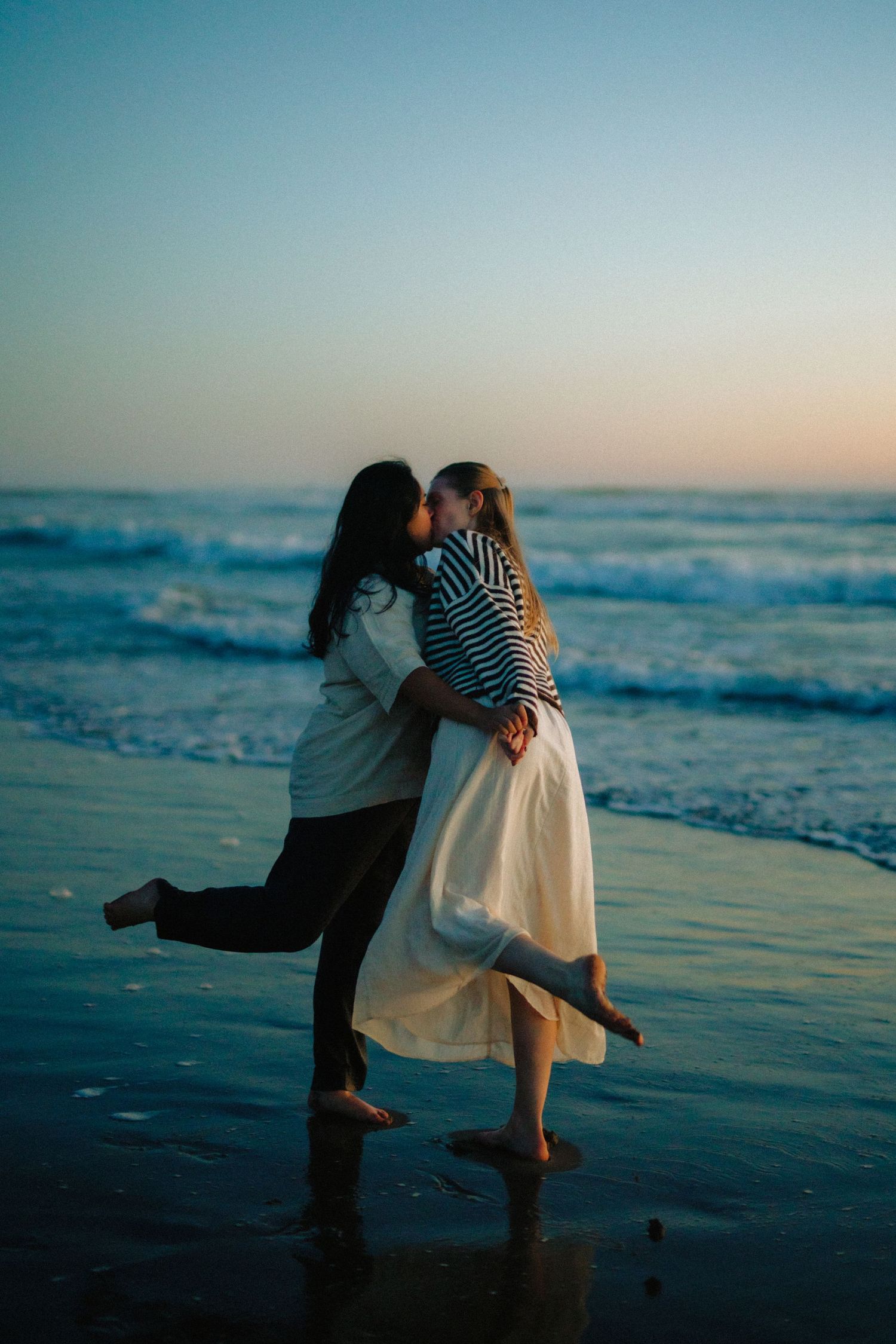Couple sharing a romantic kiss on a sandy beach at sunset, waves crashing behind them.