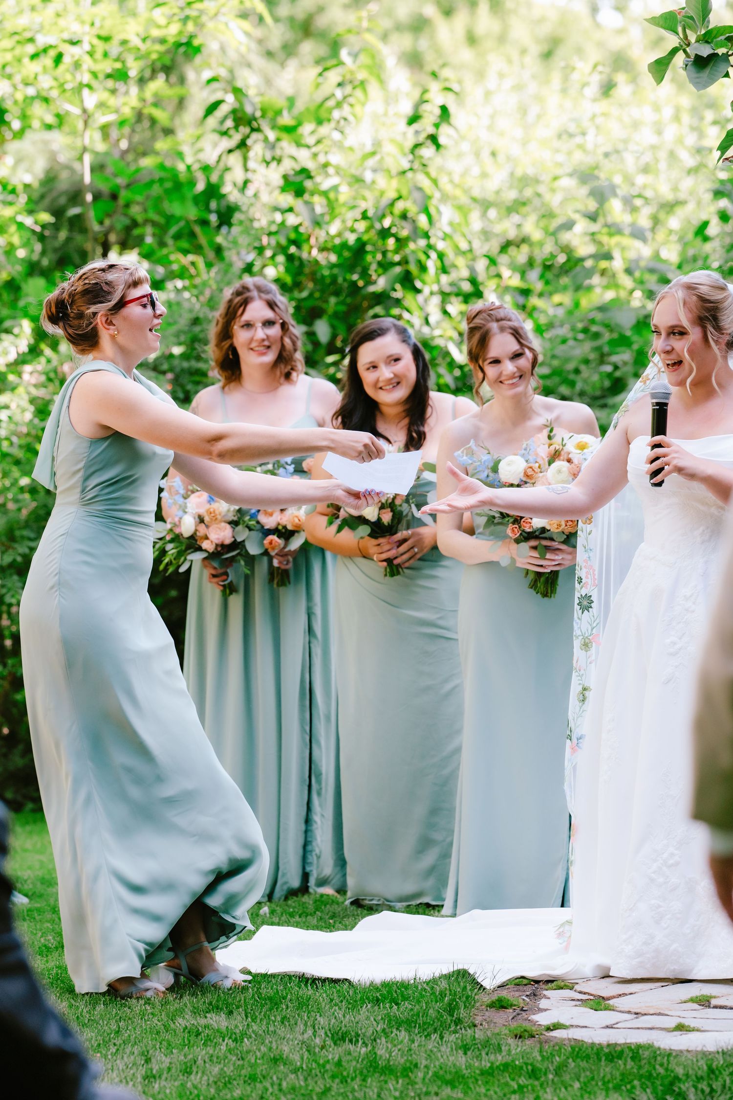 Bridesmaids in sage green dresses laugh with bride holding bouquets at outdoor wedding ceremony.