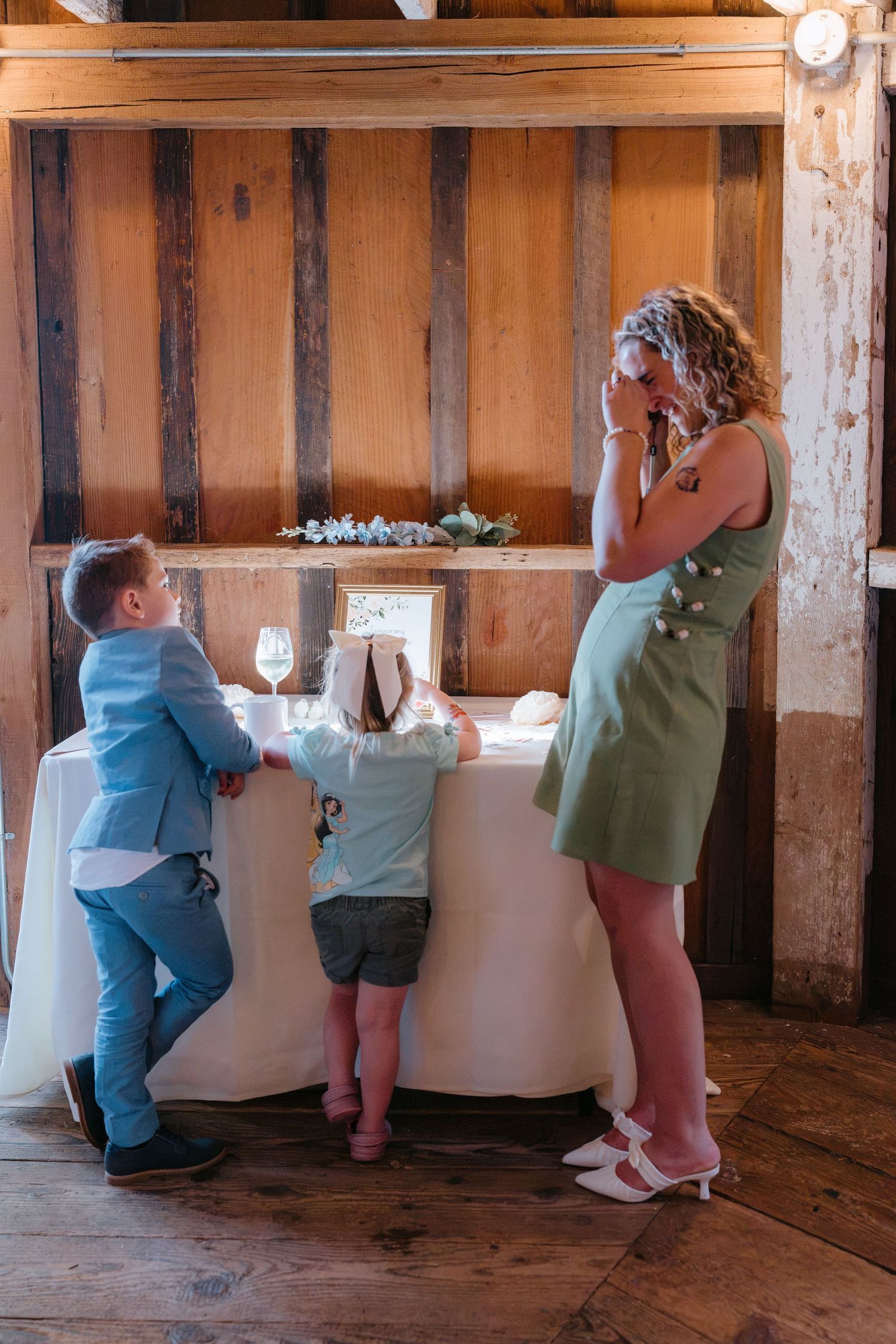 Woman in green dress with two children examining wedding guest book table in rustic barn venue.