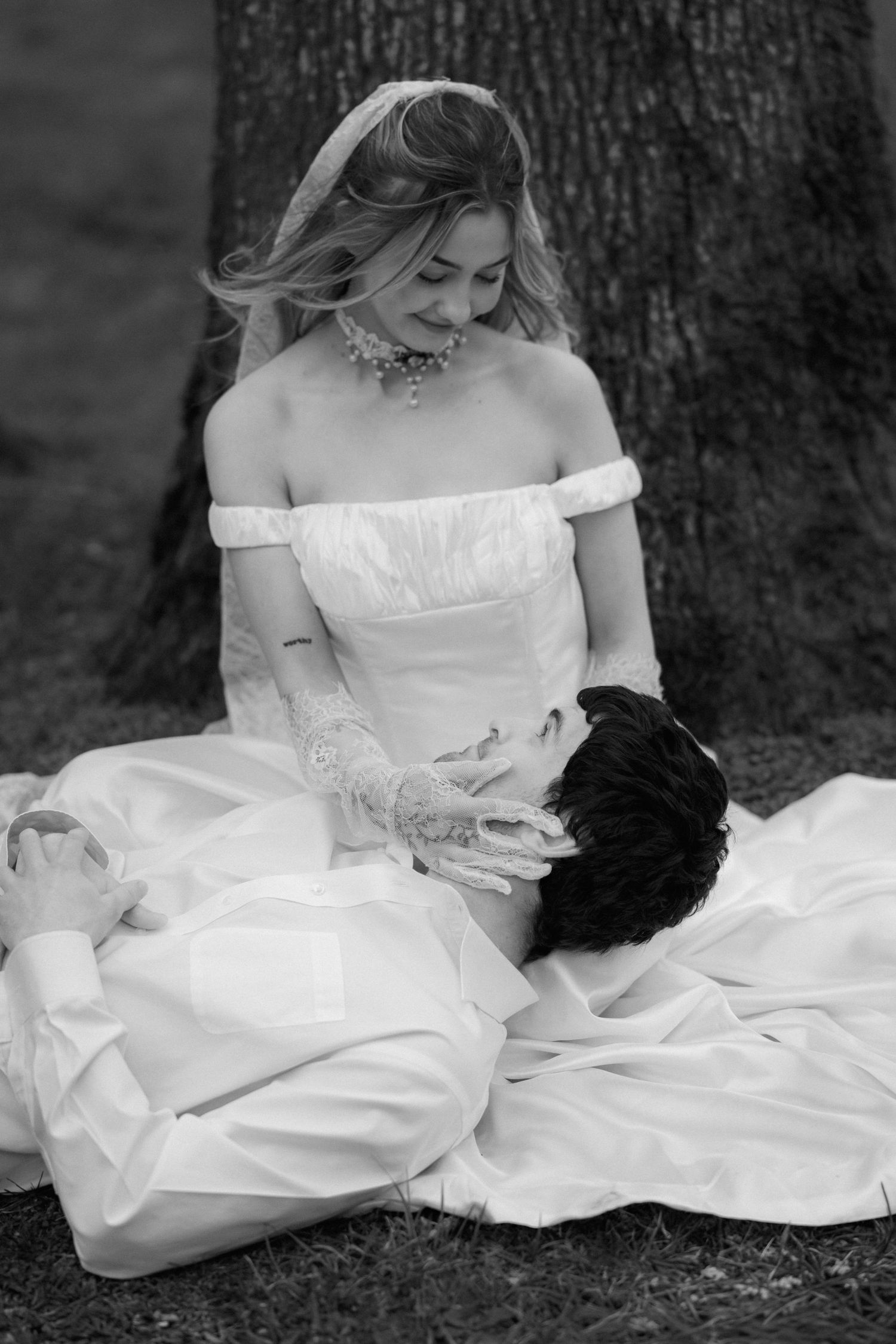 Black and white portrait of a bride in an off-shoulder wedding dress sitting gracefully on the ground.