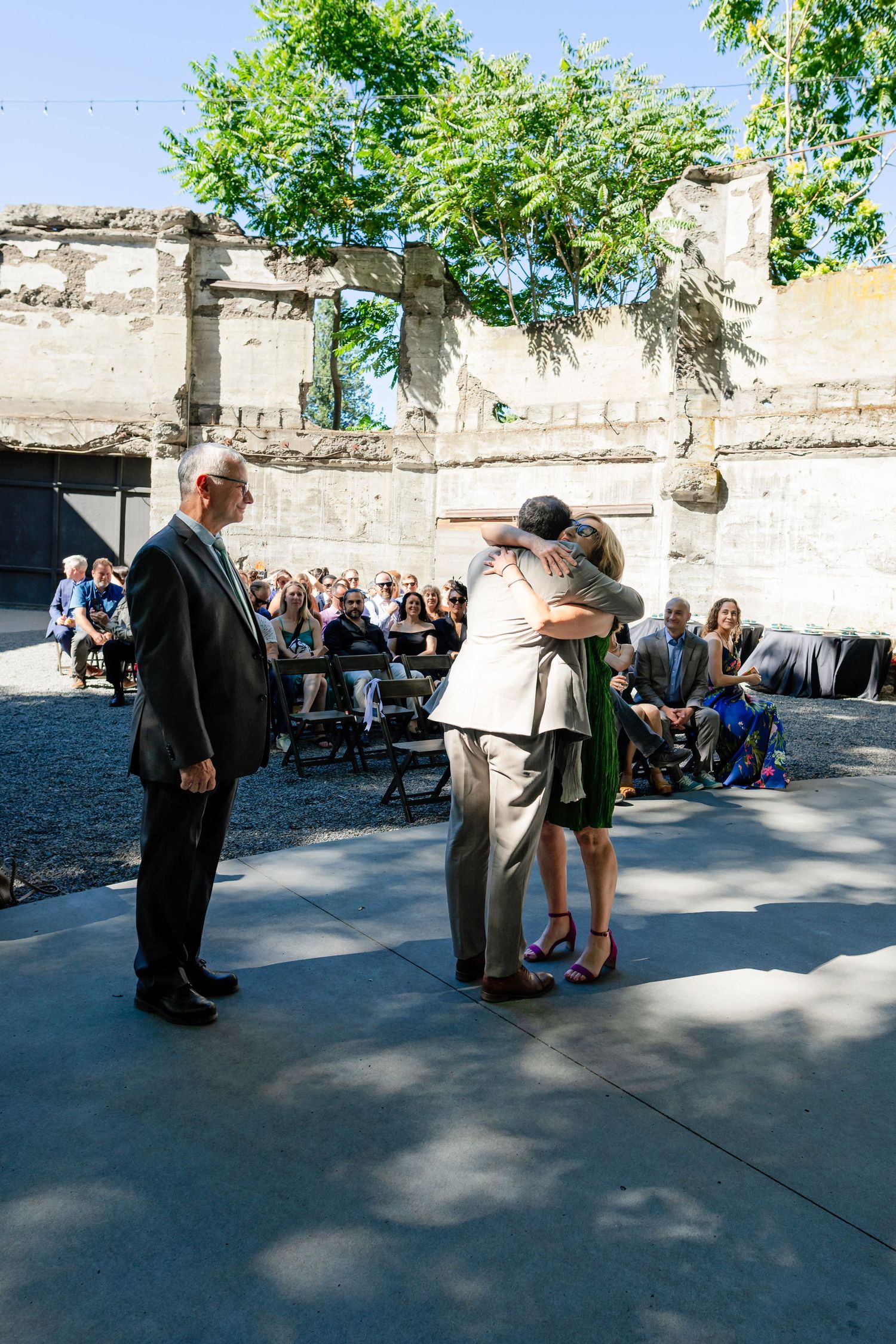 Groom embracing family member during outdoor wedding ceremony amid historic stone ruins with seated guests.