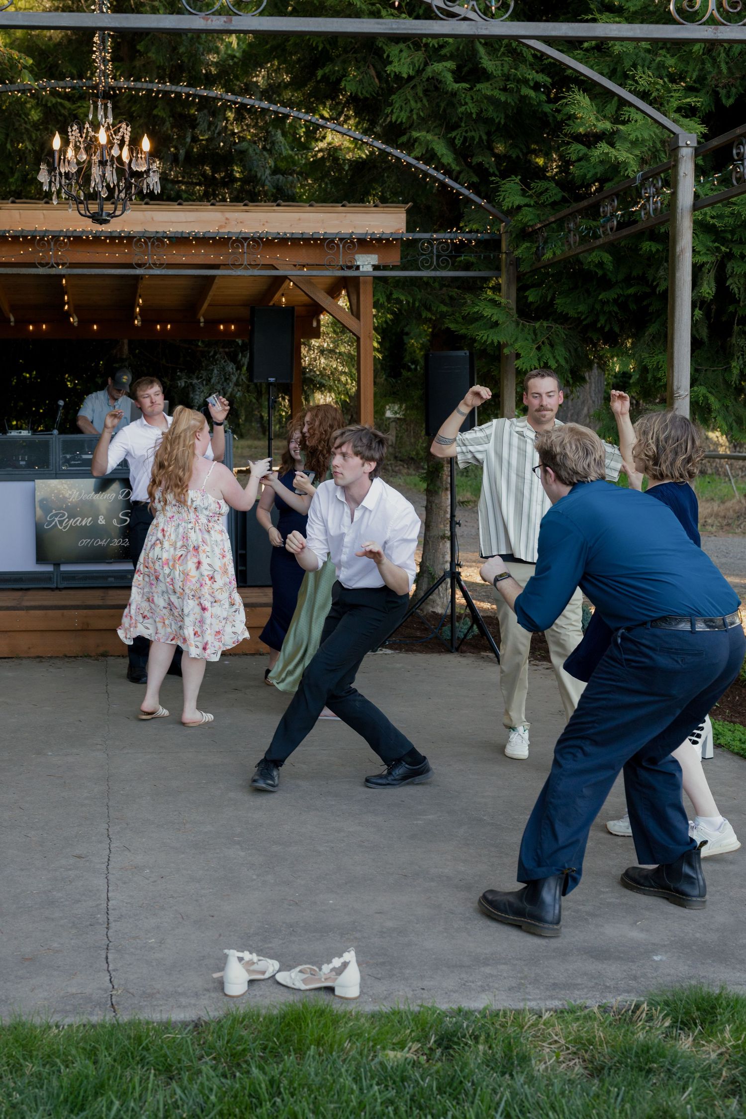 Wedding guests dance energetically on outdoor patio under chandelier and string lights at reception.