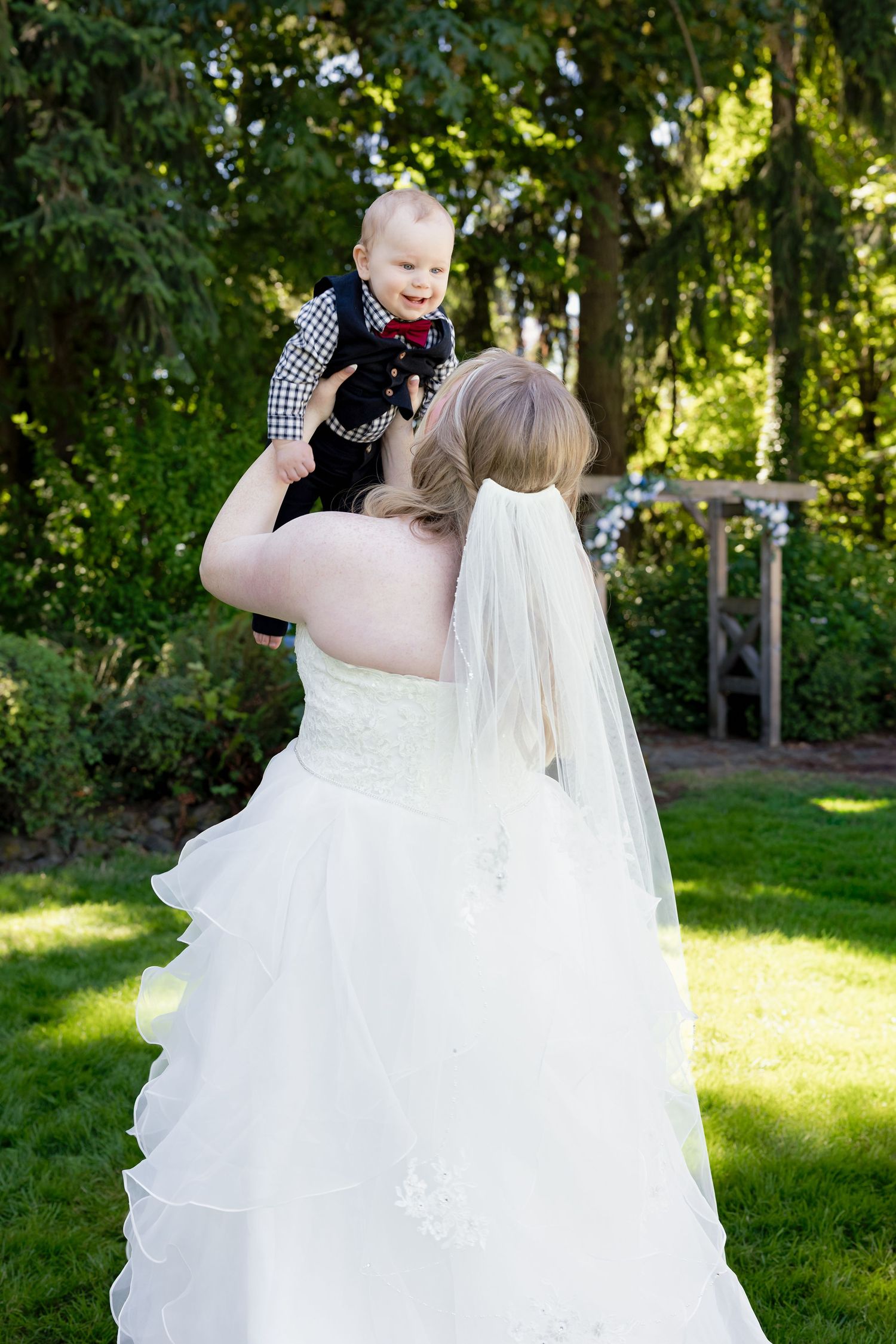 Bride in white gown and veil lifting a smiling baby boy in a black outfit at an outdoor wedding.