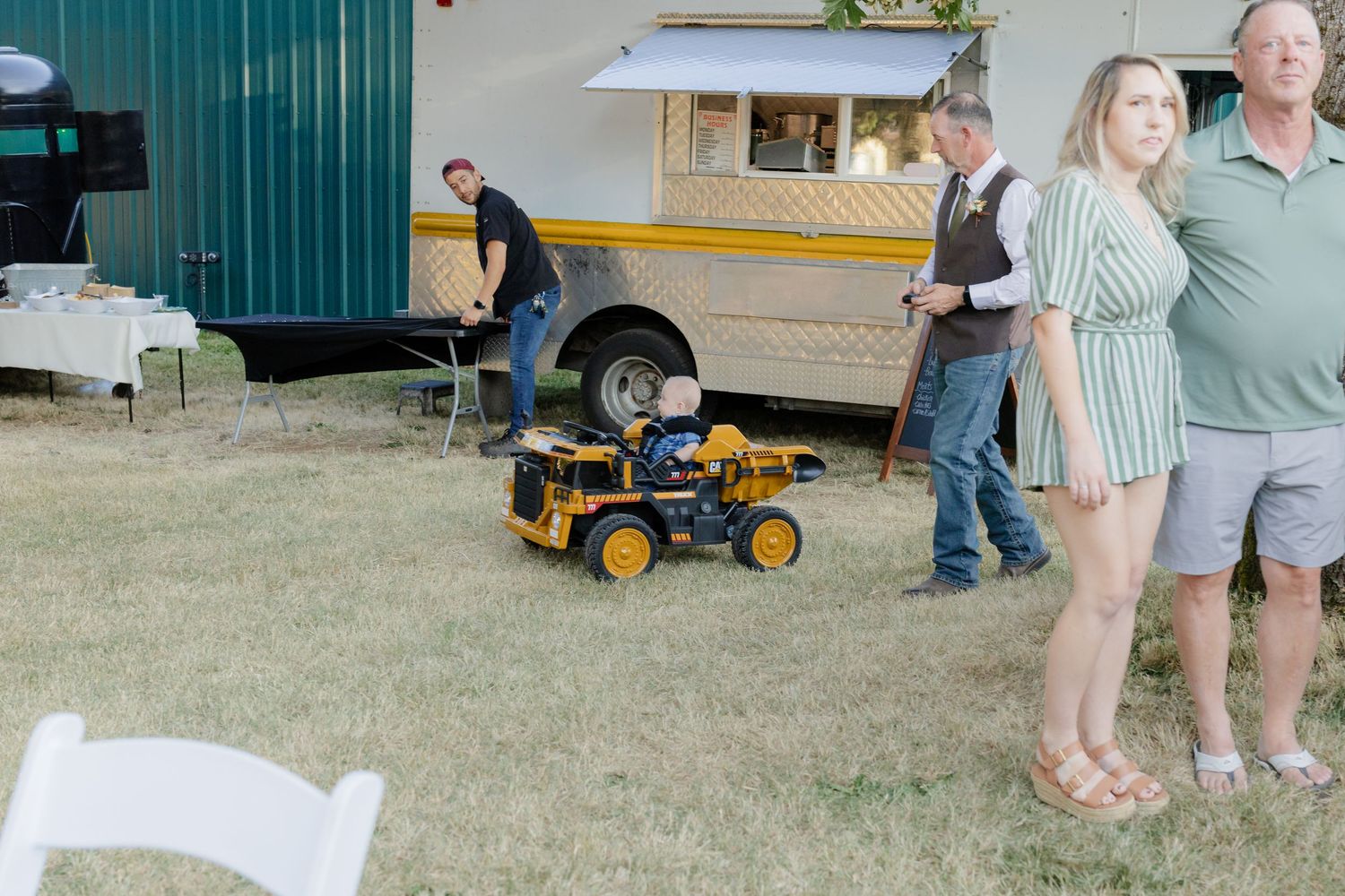 Child riding a toy yellow bulldozer at an outdoor wedding venue near a food truck with guests nearby.