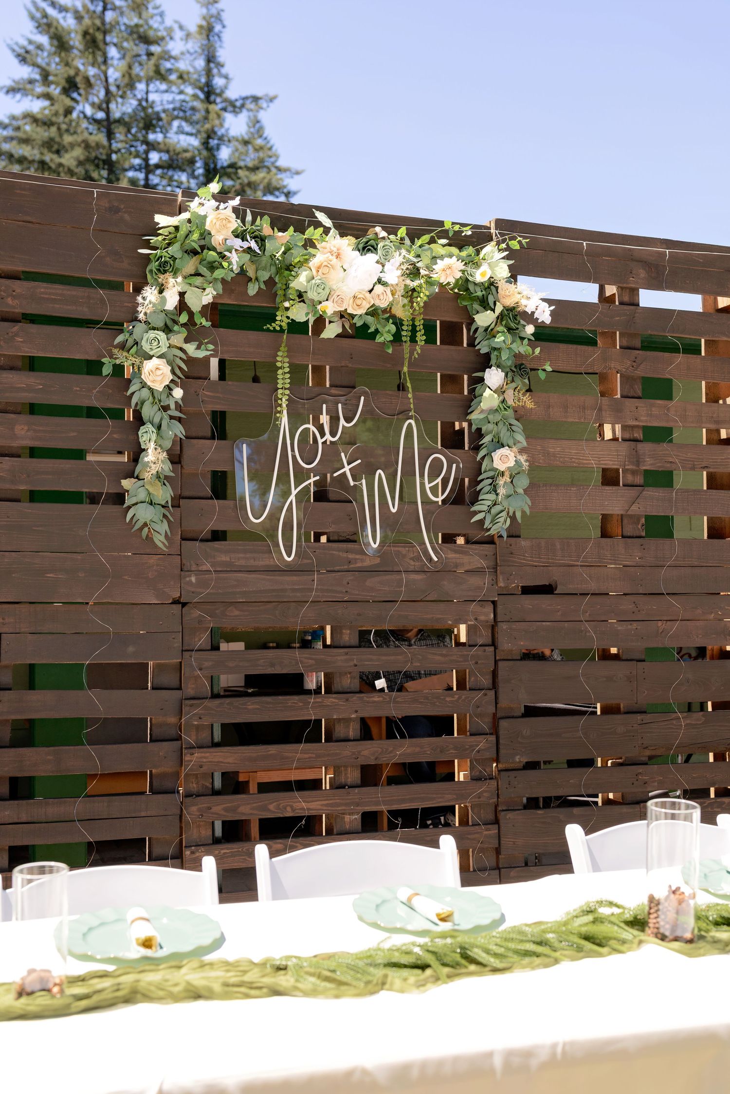 Rustic wooden pallet wedding backdrop with floral arch and neon You & Me sign above an elegant outdoor reception table.