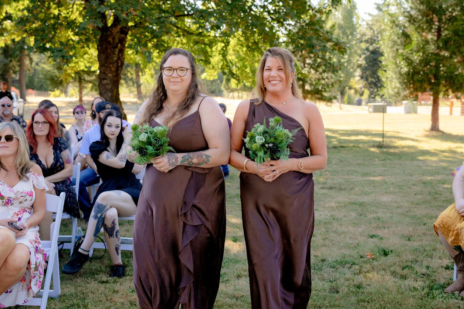 Two bridesmaids in brown one-shoulder dresses walking down outdoor wedding aisle holding green bouquets.