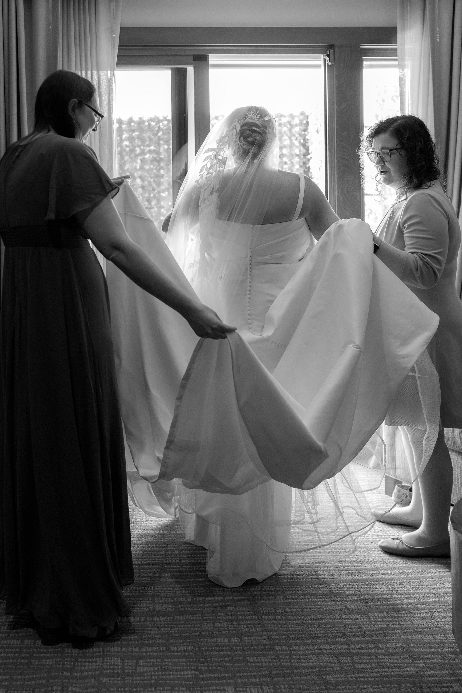Silhouetted figures help adjust flowing wedding dress train in sunlit room.