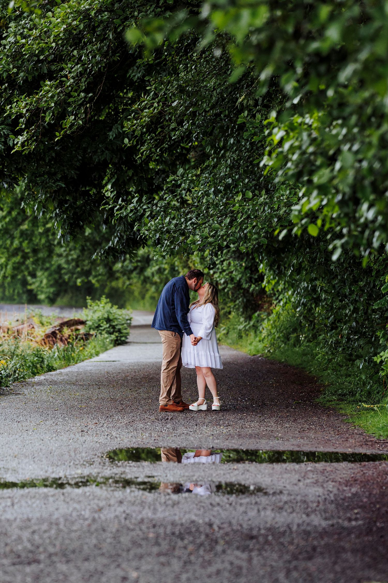 A couple embraces on a wet path surrounded by lush green foliage and trees in a romantic garden setting.