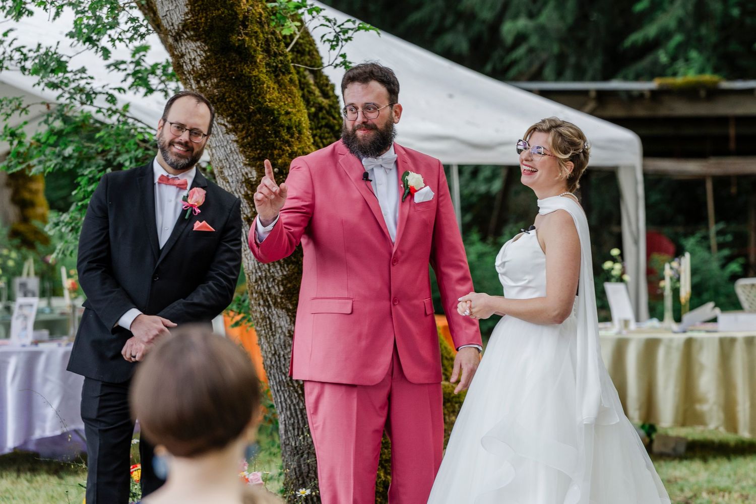 An outdoor wedding ceremony under trees with formal attire and a distinctive pink suit.