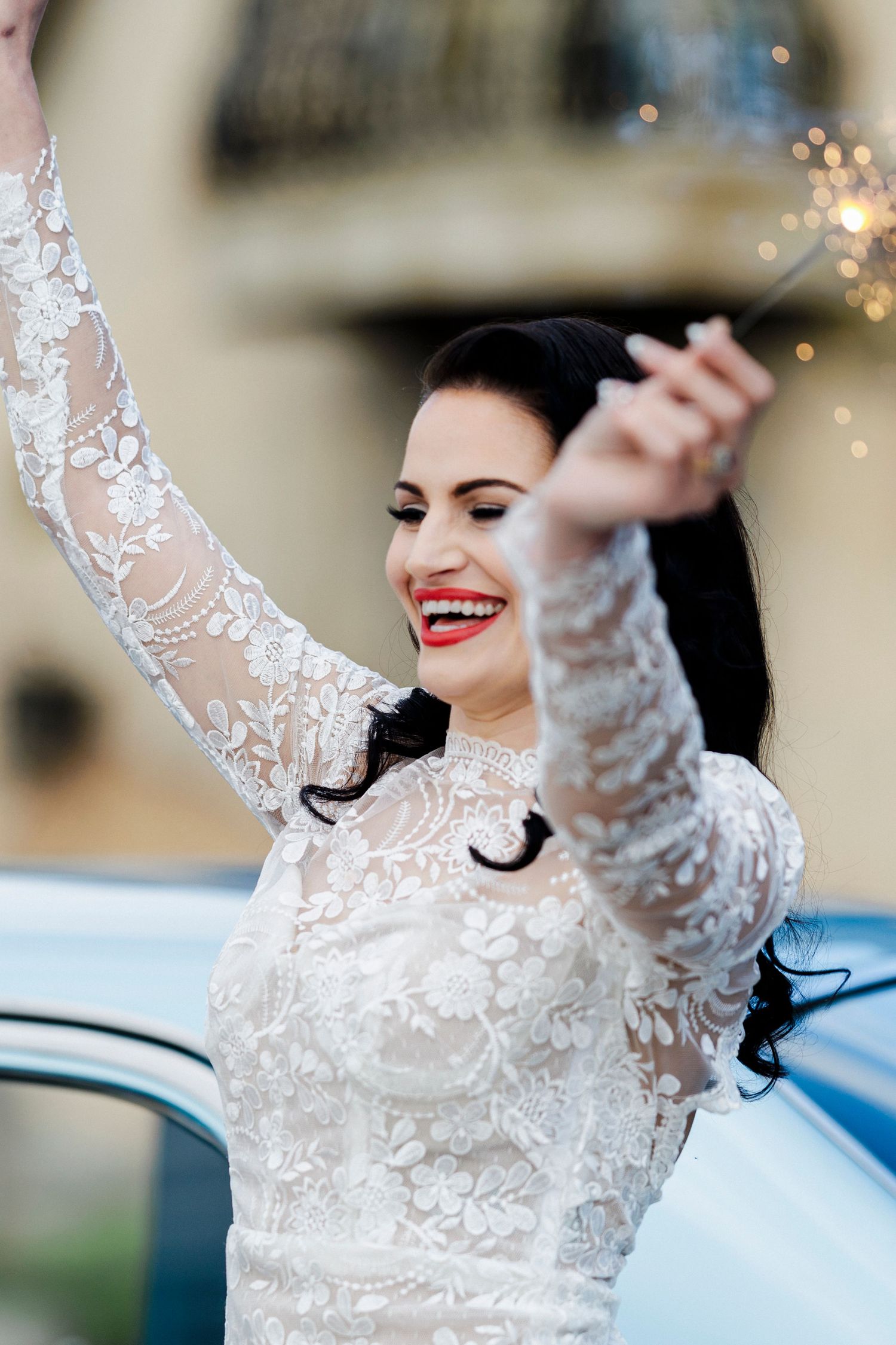 A joyful bride in a long-sleeved lace wedding dress smiles with bright red lipstick while raising one arm in celebration.
