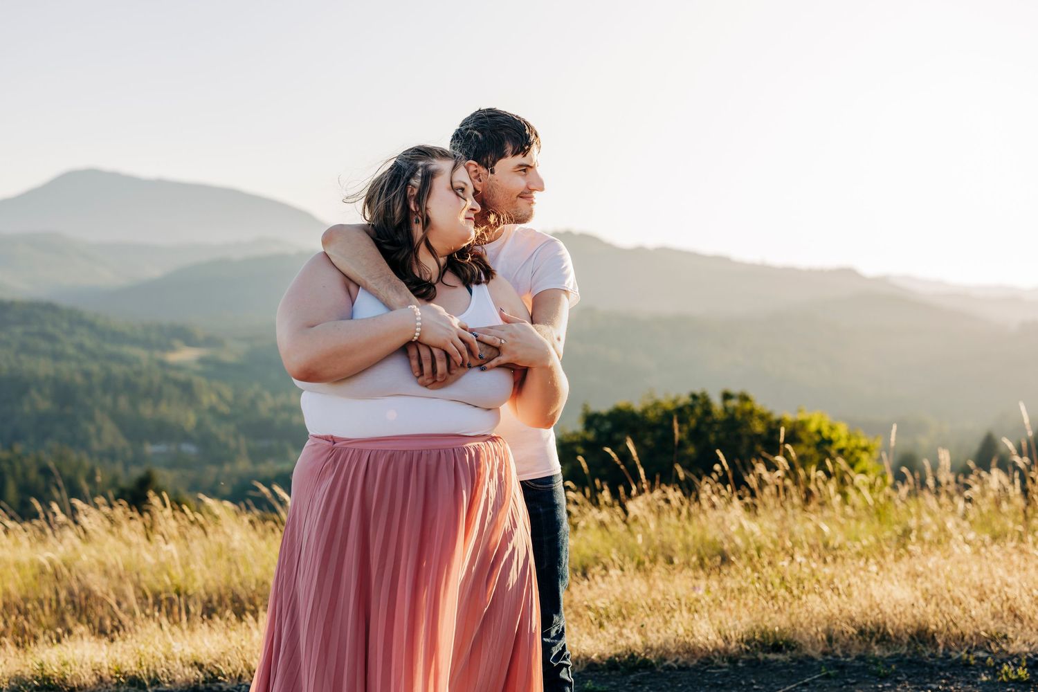 A couple embraces while standing in a golden field at sunset with mountains in the background.