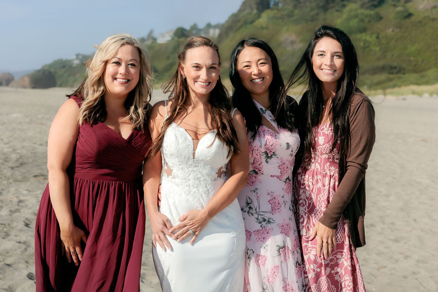 A group of wedding attendants in burgundy and floral dresses pose together on a scenic beach with mountains in background.