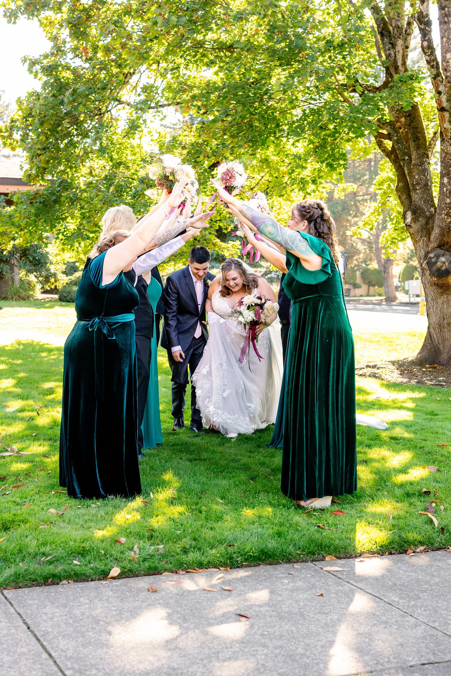 Wedding party in emerald green velvet dresses forming archway outdoors.
