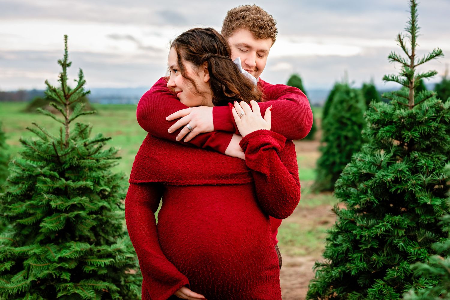 A couple in red sweaters embraces among evergreen trees at a Christmas tree farm during golden hour.