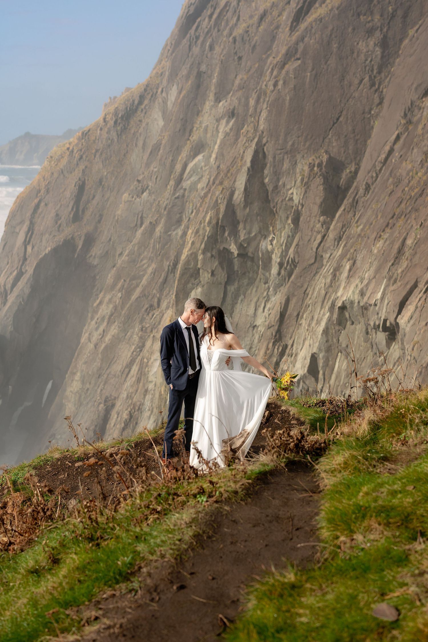 A couple in wedding attire shares an intimate moment on a dramatic coastal cliff overlooking the ocean at sunset.