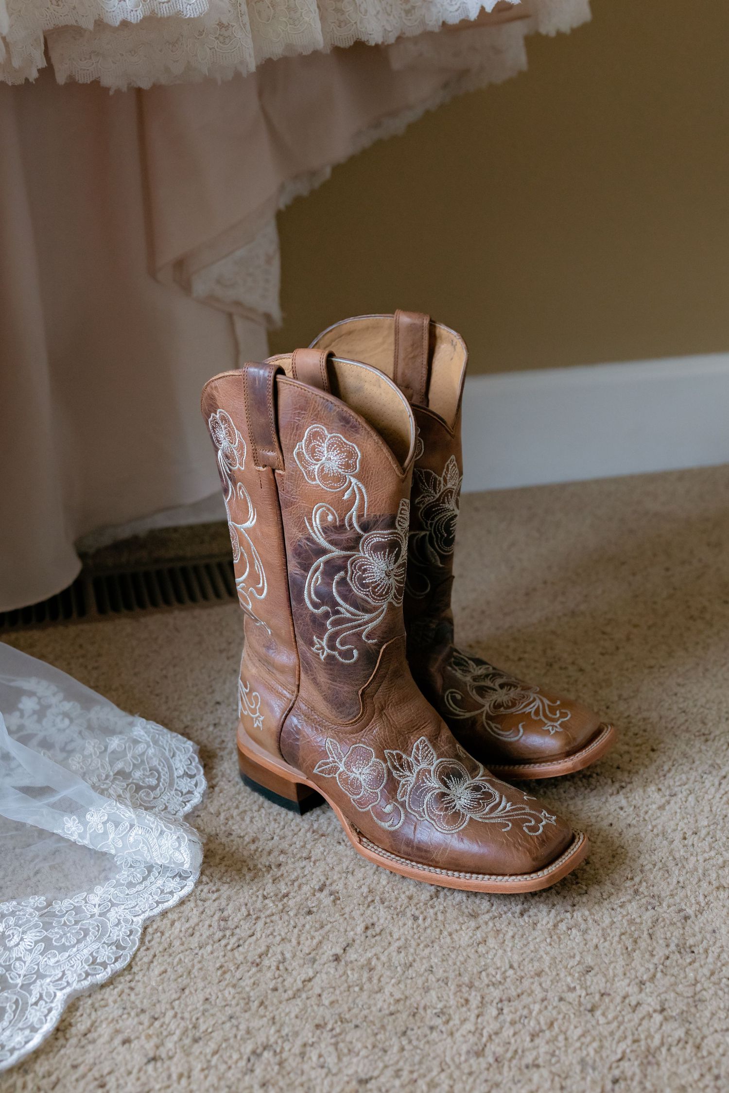 Brown leather cowboy boots with floral embroidery displayed on carpet beside a lace wedding dress.