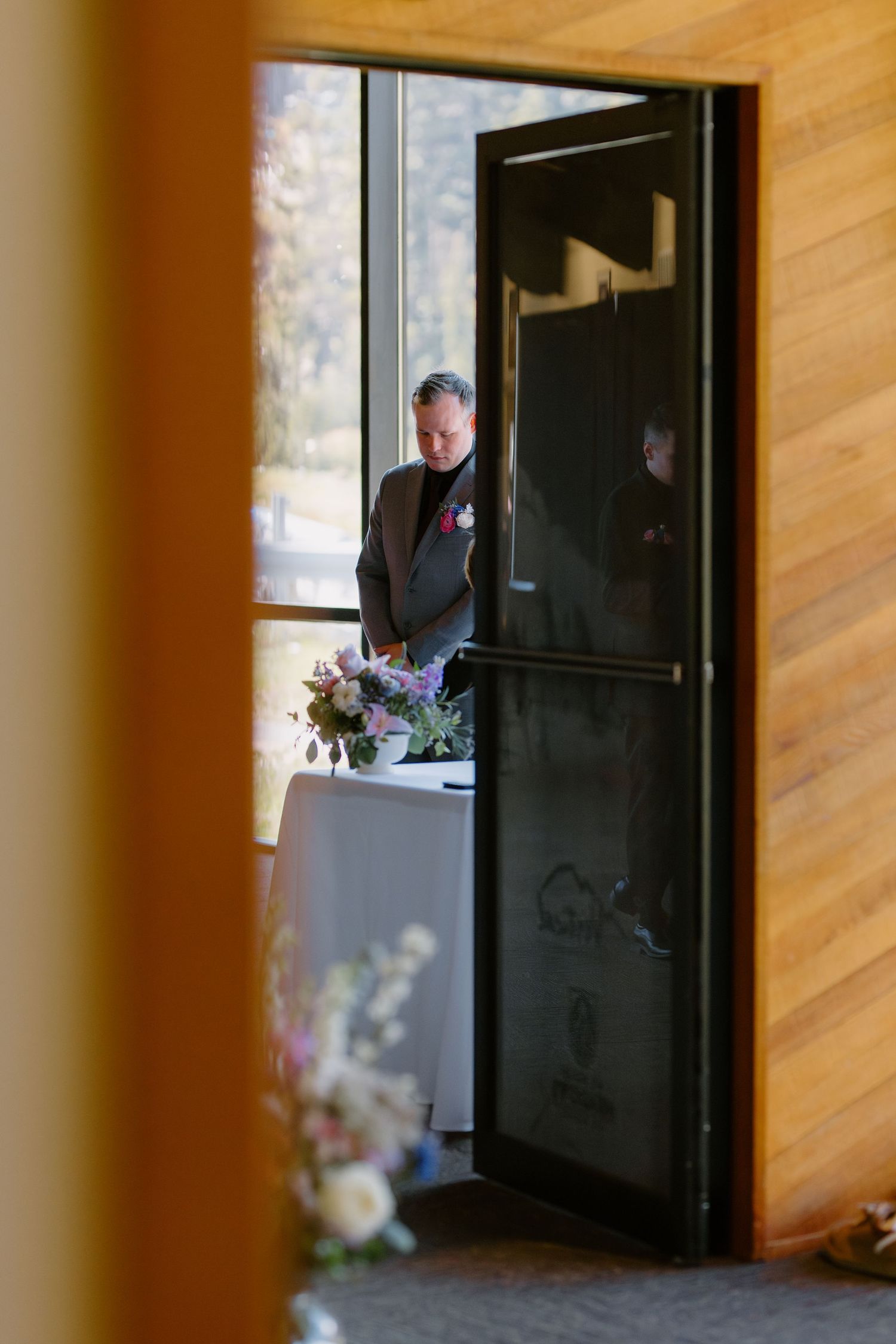 Groom in brown suit standing at floral arrangement table viewed through open doorway of wood-paneled venue.